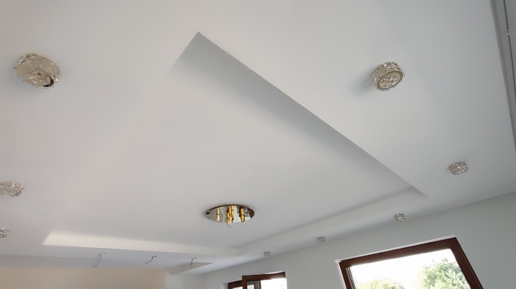 Biały sufit podwieszany z dekoracyjnym wgłębieniem i punktowymi lampami w stylu glamour. Widoczne okno i fragment ściany. Minimalistyczny design z nutą elegancji.