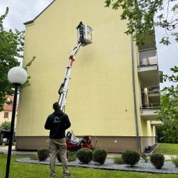 Kopacek - Mycie elewacji budynku z podnośnika koszowego. Widoczny pracownik w koszu i osoba nadzorująca z dołu. Żółta elewacja z widocznymi smugami wody.