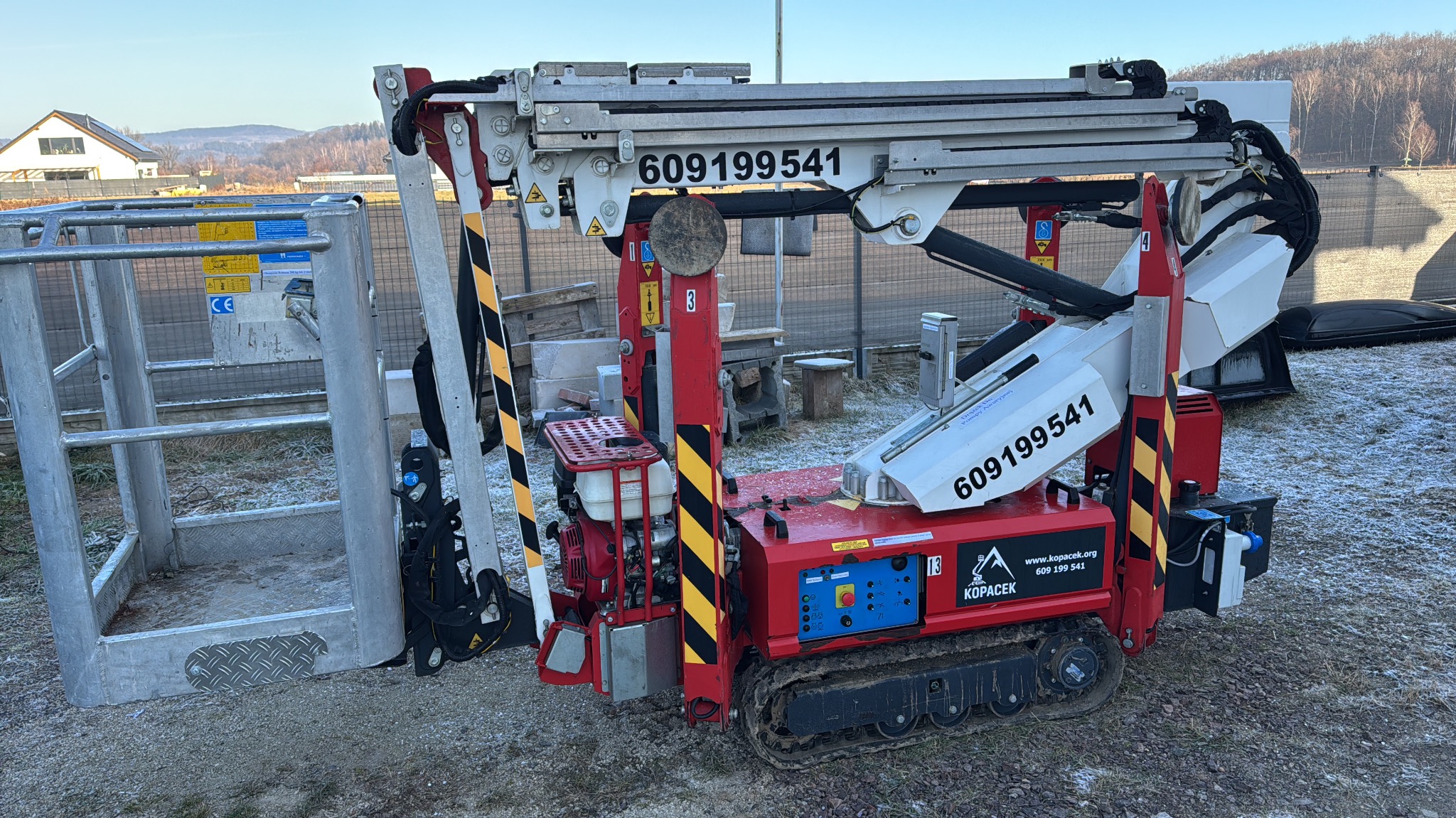 Czerwono-biała mini-dźwig gąsienicowy KOPACEK z numerem telefonu 609199541 na tle krajobrazu z domem i drzewami. Widoczny kosz podnośnika.