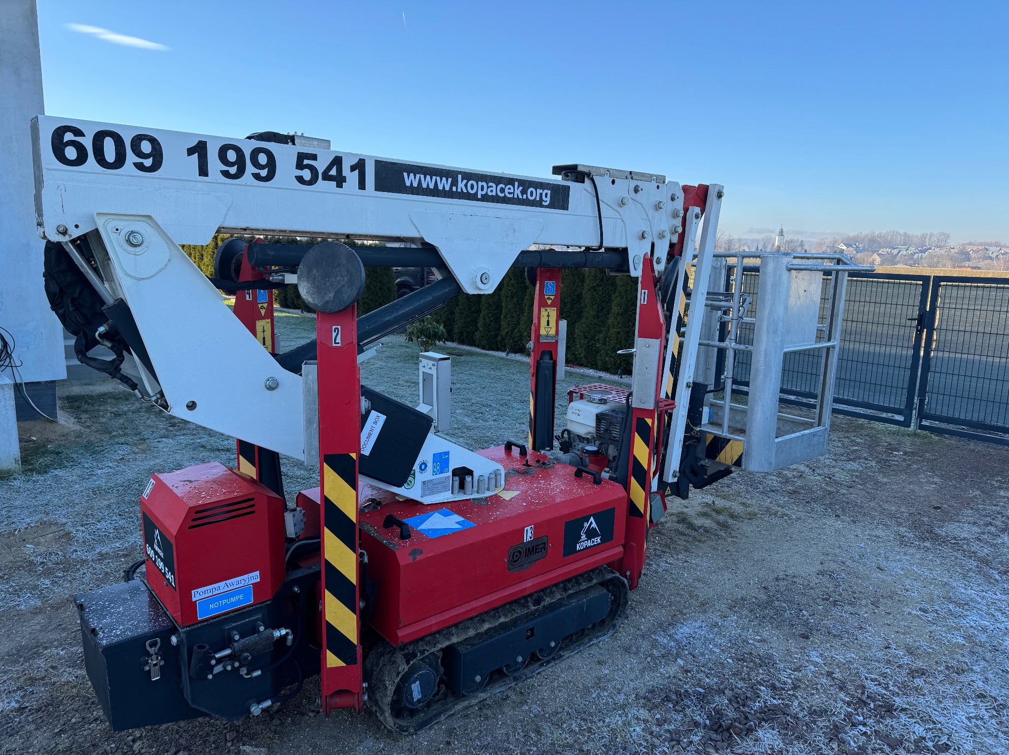 Podnośnik koszowy gąsienicowy z numerem telefonu i adresem strony internetowej na ramieniu, zaparkowany na zewnątrz w chłodny dzień.