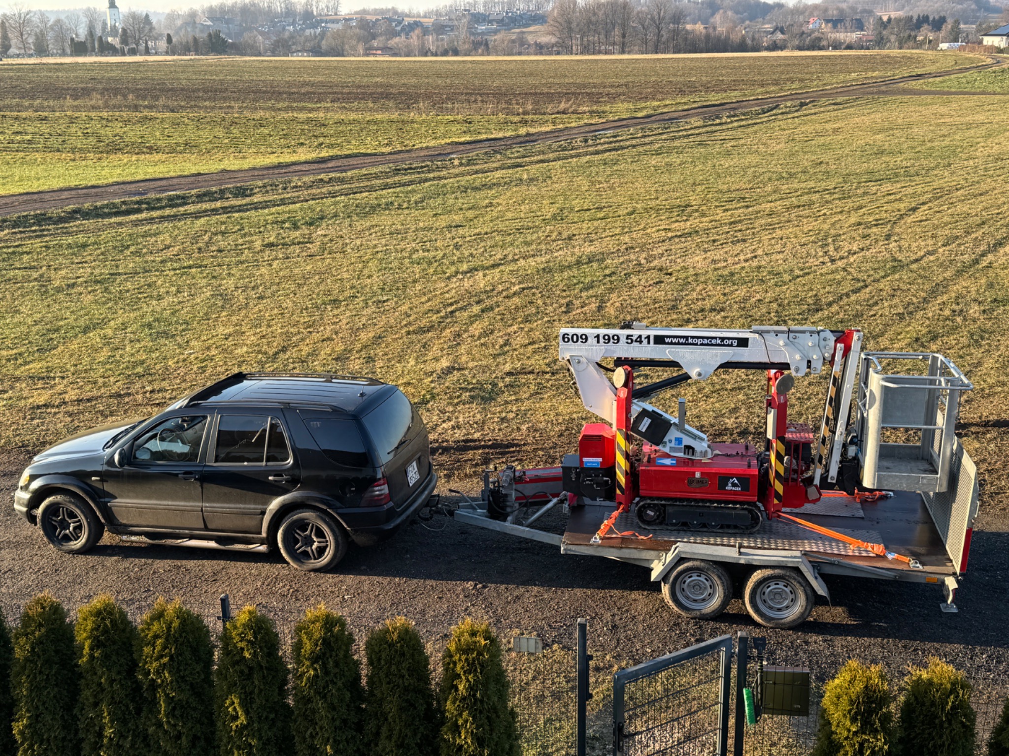 Czarny SUV ciągnie przyczepę z podnośnikiem koszowym na tle pola i zabudowań. Widoczne tuje wzdłuż ogrodzenia.