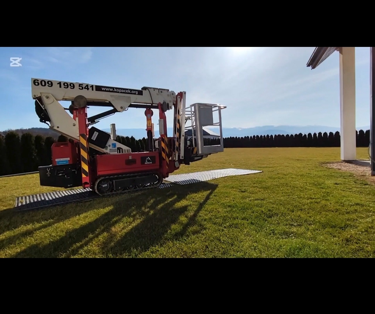 Czerwony podnośnik koszowy gąsienicowy na trawniku, z widocznym koszem i wysięgnikiem, na tle błękitnego nieba i gór. Widoczny numer telefonu i adres strony internetowej.