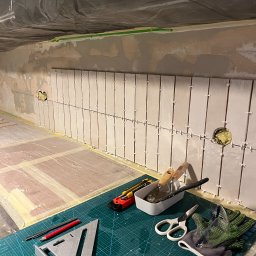 Wiktor Lipski - Montaż białych, prostokątnych płytek na ścianie w kuchni, w trakcie pracy. Widoczne narzędzia: poziomica, szpachelki, nożyczki, rękawice ochronne i zapas płytek.