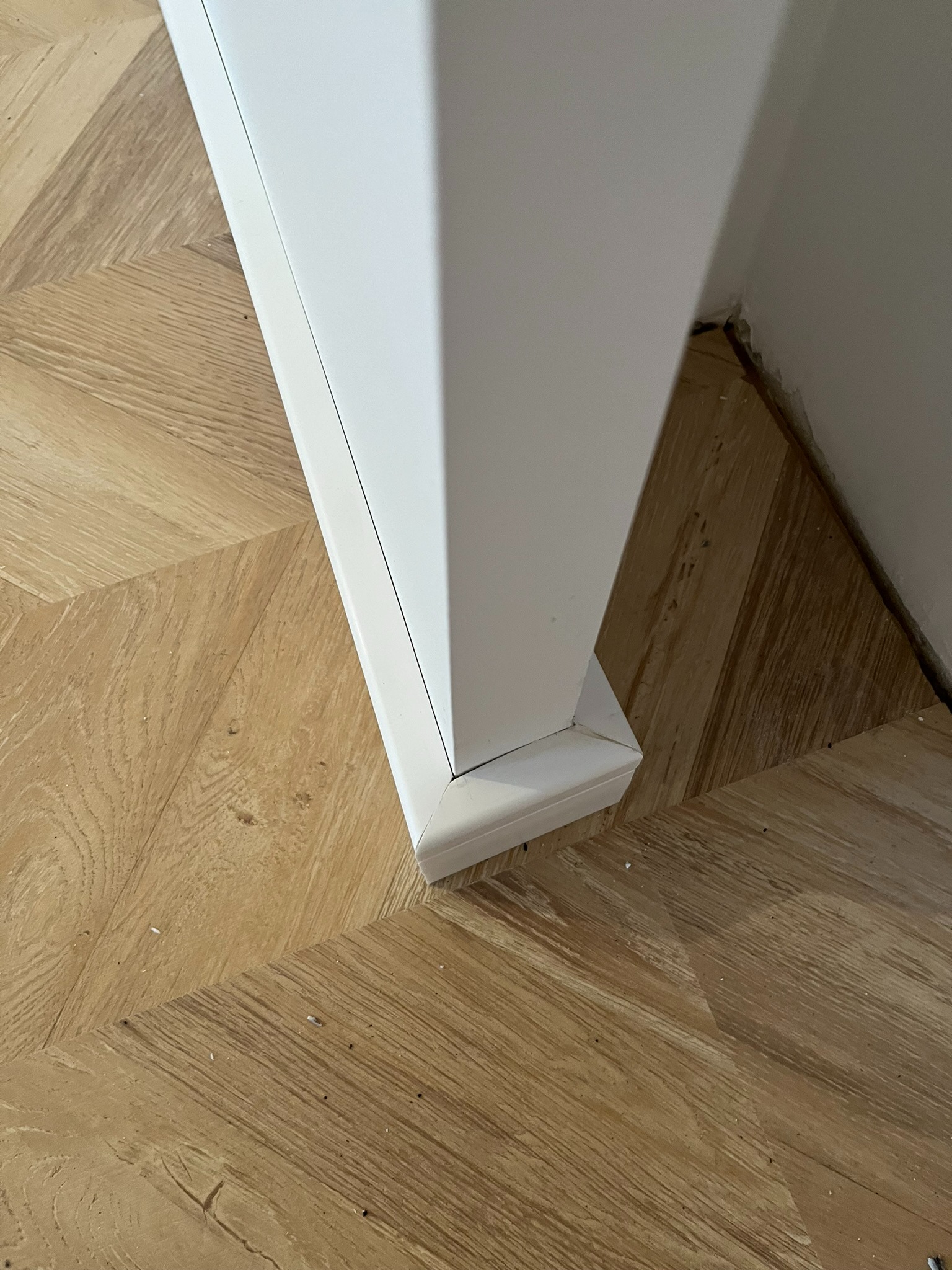 Detal montażu białej listwy przypodłogowej przy ścianie i parkiecie ułożonym we wzór jodełki francuskiej. Widoczne drobne zabrudzenia na podłodze.
