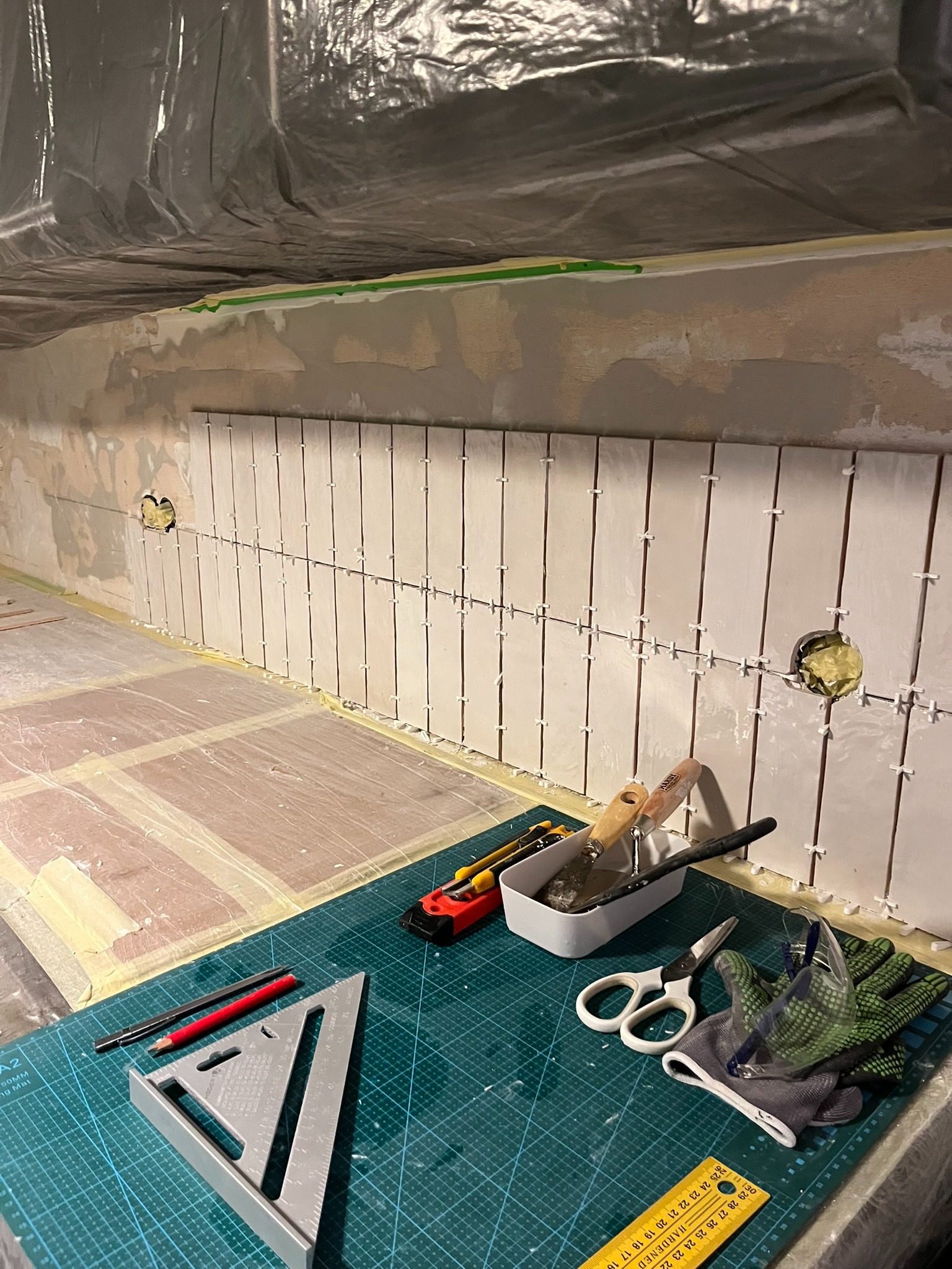 Montaż białych, prostokątnych płytek na ścianie w kuchni, w trakcie pracy. Widoczne narzędzia: poziomica, szpachelki, nożyczki, rękawice ochronne i zapas płytek.