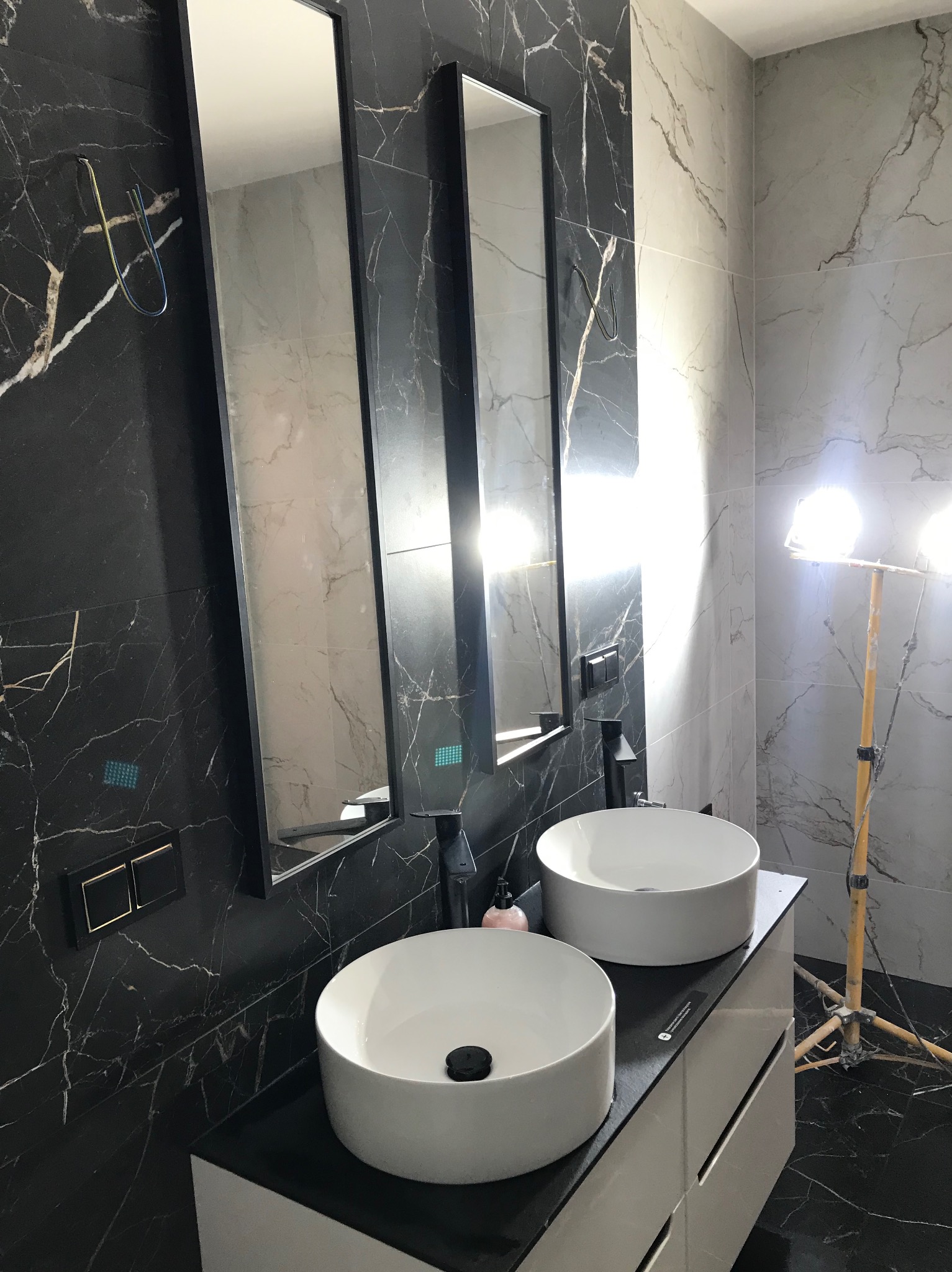 Wykończona łazienka z dwoma umywalkami nablatowymi, czarnymi bateriami i lustrami w czarnych ramach na tle marmurowych płytek. Widoczne oświetlenie robocze.