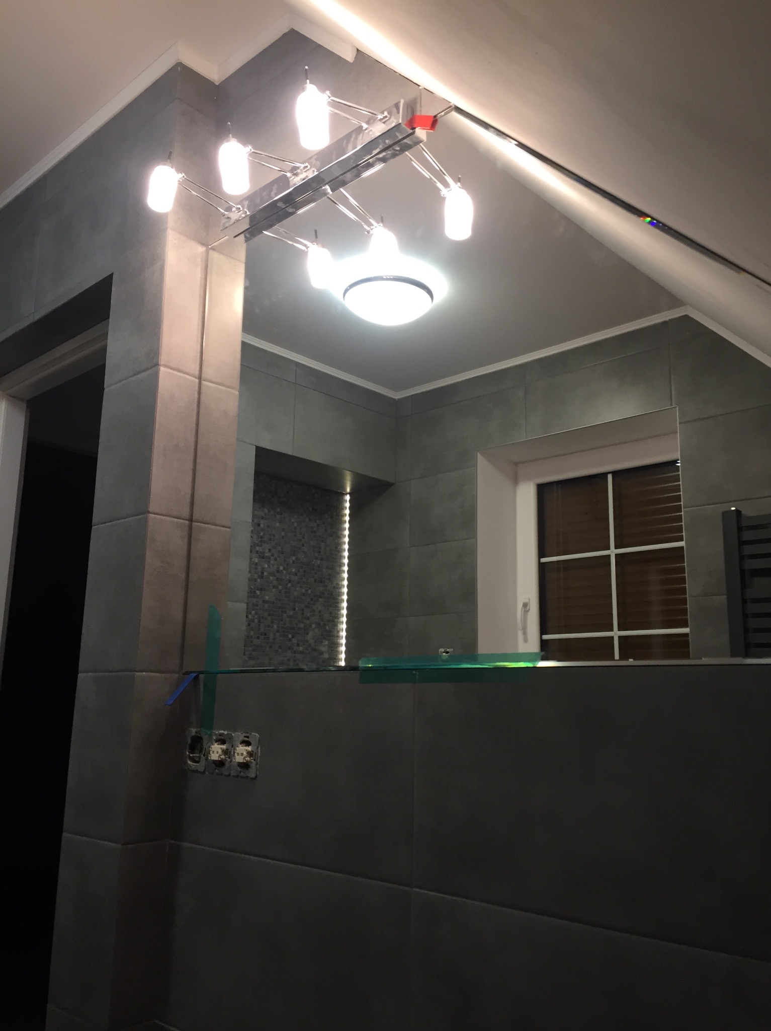 Nowoczesna łazienka z szarymi płytkami, oświetleniem punktowym i lustrem. Widoczne gniazdka elektryczne bez osprzętu. Ukończone wykończenie wnętrza.