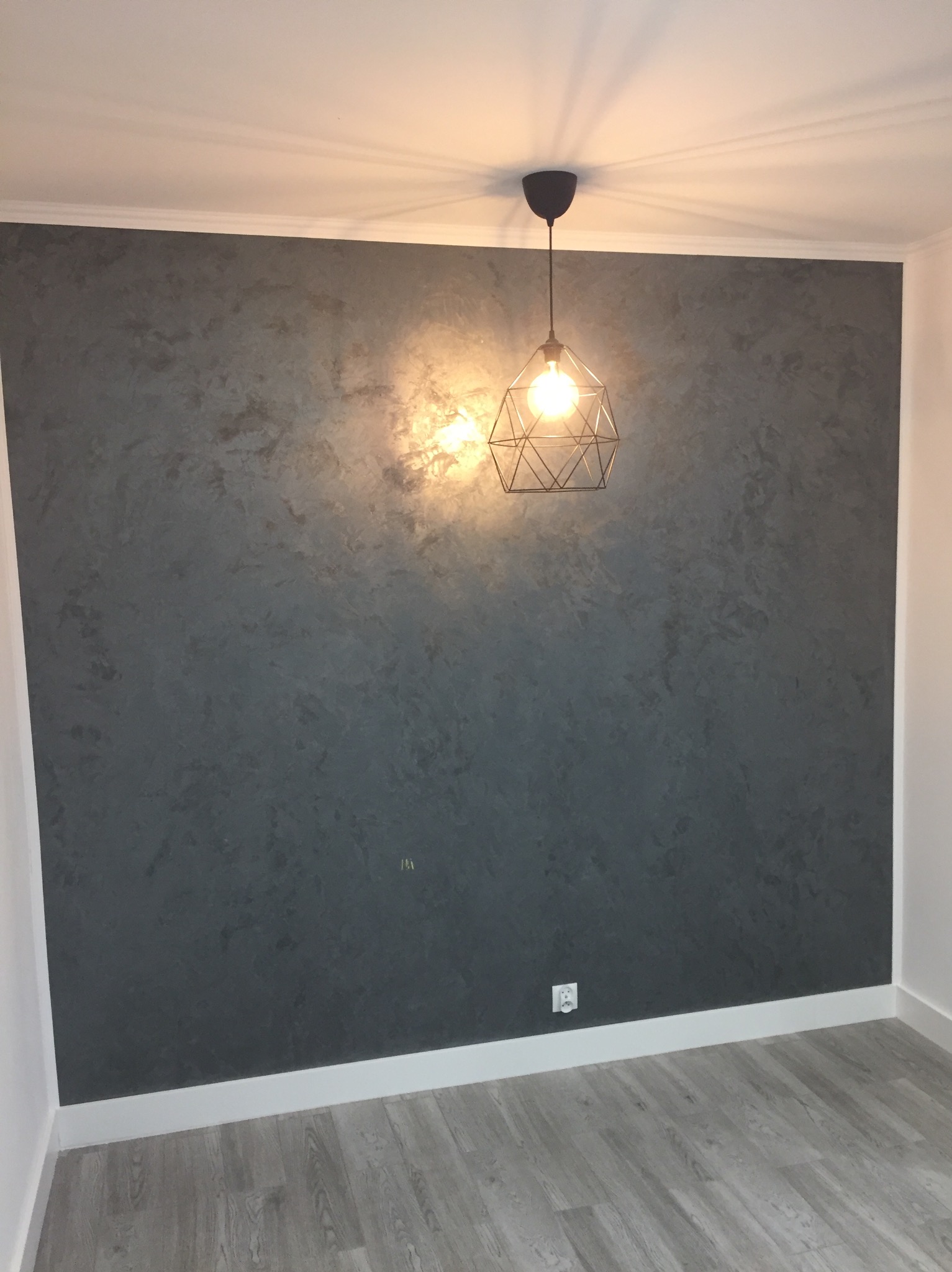 Ściana z efektem dekoracyjnym w szarościach, oświetlona lampą w Wołominie. Nowoczesne wykończenie wnętrza z widoczną strukturą i gładką podłogą.