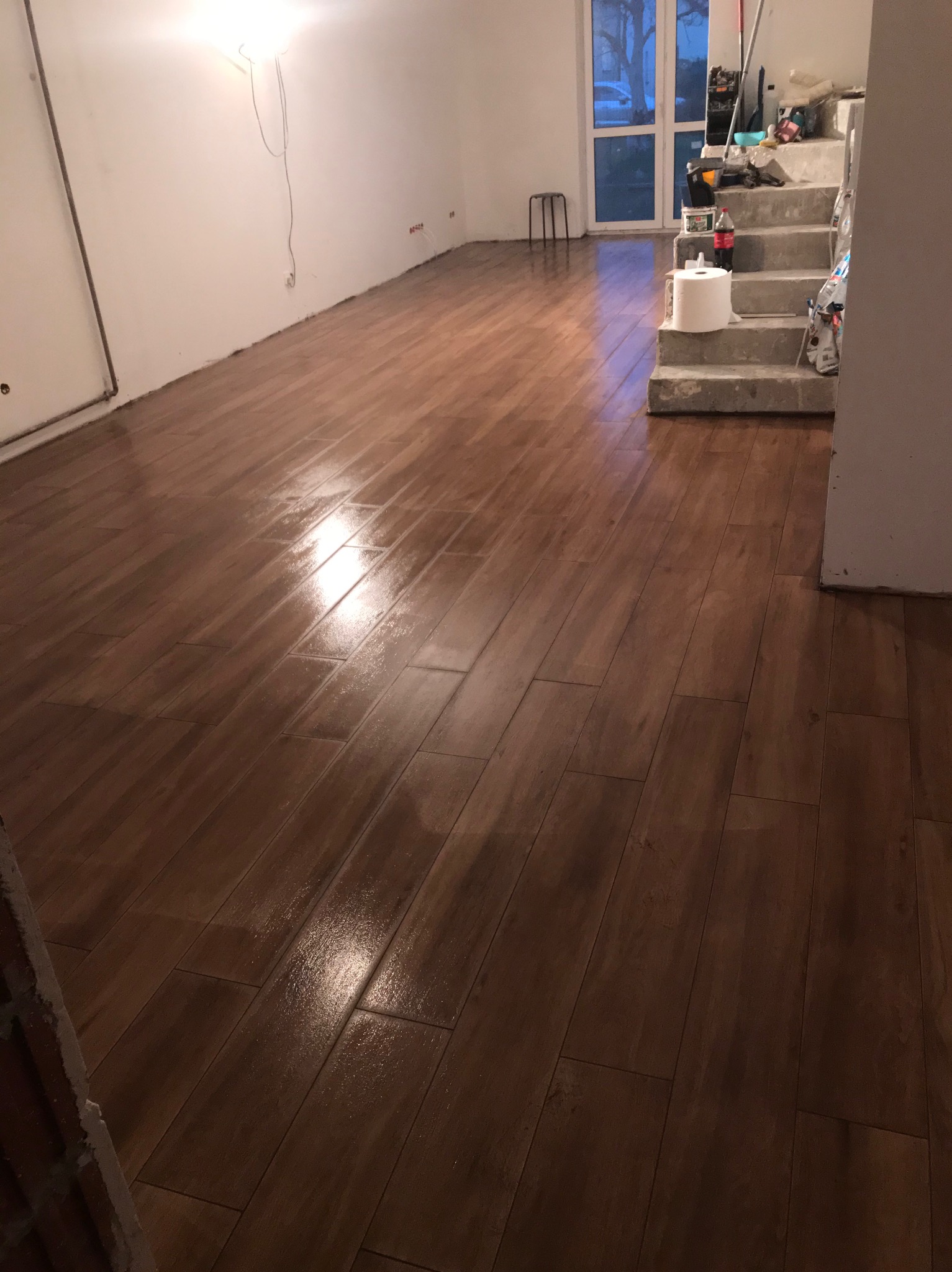 Ukończona podłoga z brązowych płytek imitujących drewno w jasnym pomieszczeniu. Widoczne schody w tle i narzędzia, co sugeruje trwające prace wykończeniowe.