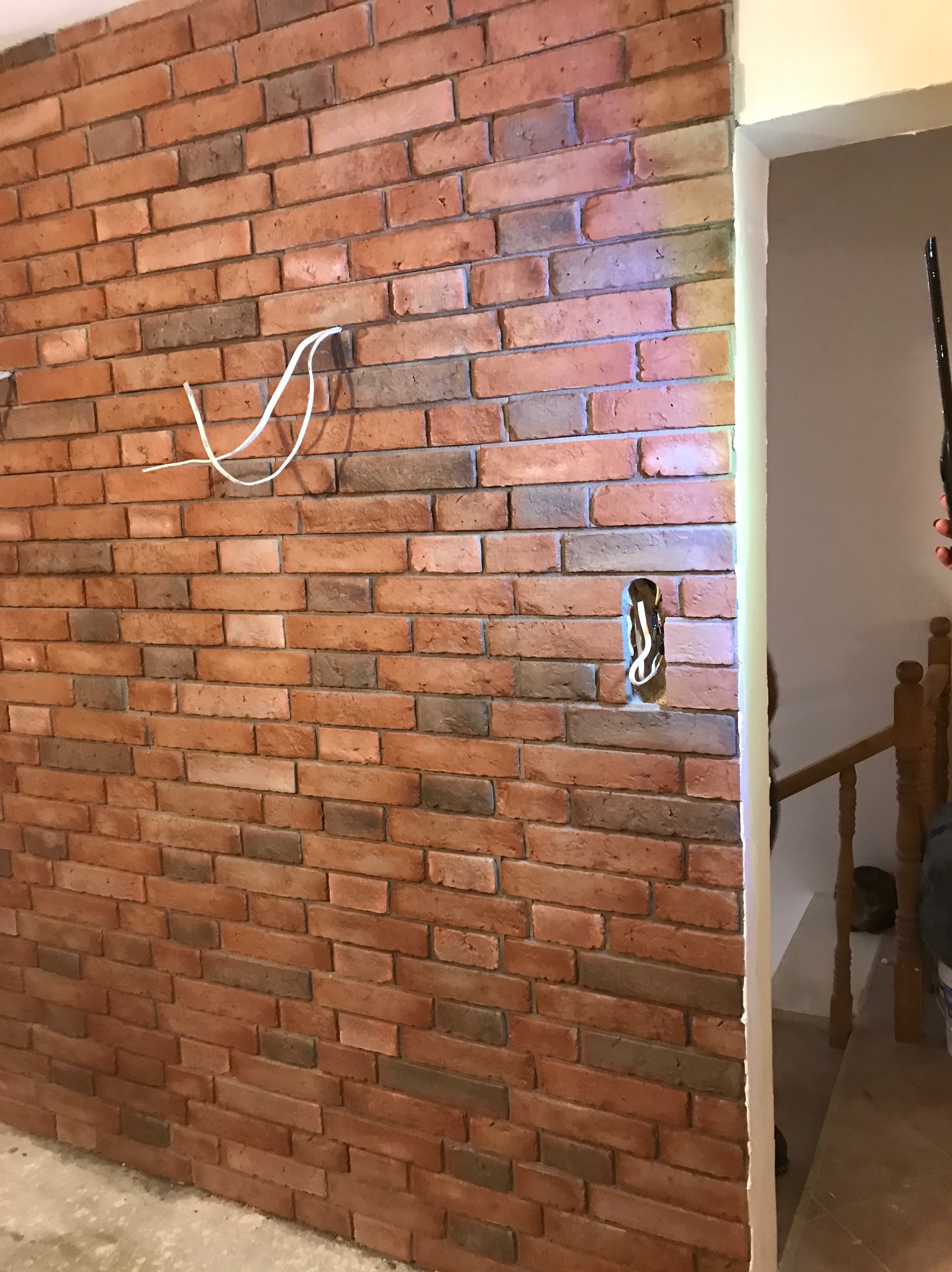 Ściana z cegły klinkierowej w odcieniach czerwieni i brązu, z widocznymi przewodami elektrycznymi i fragmentem korytarza z drewnianą balustradą w tle.