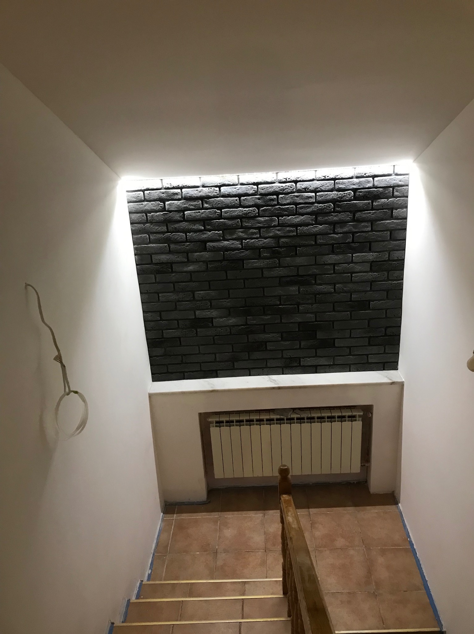 Klatka schodowa z dekoracyjną ścianą z czarnej cegły, podświetloną od góry. Widoczny grzejnik i fragment schodów z drewnianą barierką. Wykończenie w stylu nowoczesnym.