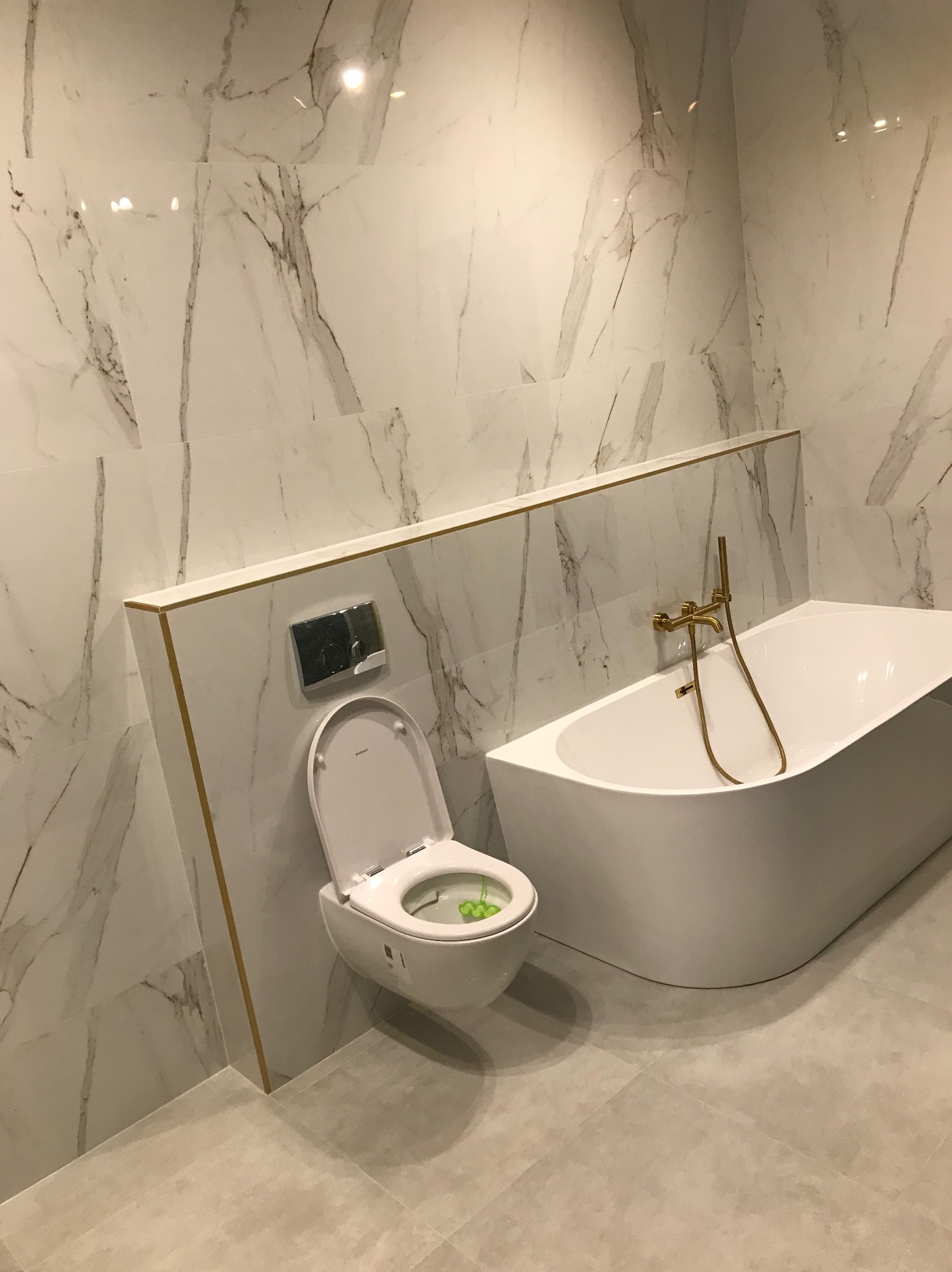 Nowoczesna łazienka z marmurowymi płytkami, białą wanną i toaletą. Złota armatura dodaje elegancji. Minimalistyczny design i jasne kolory tworzą przestronne wnętrze.