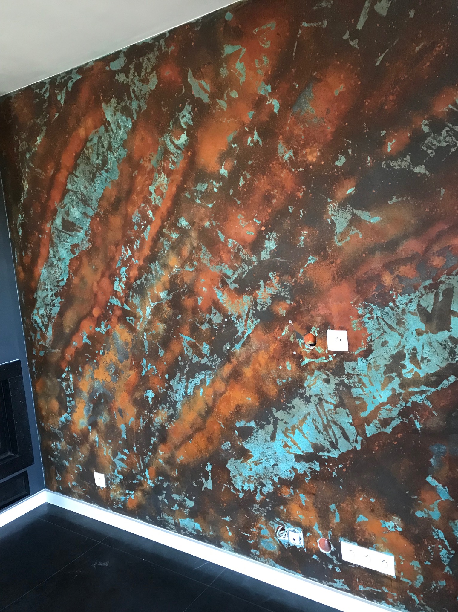 Ściana z dekoracyjną fakturą w odcieniach miedzi i turkusu, widoczne gniazdka elektryczne. Unikalne wykończenie wnętrza z efektem patyny.