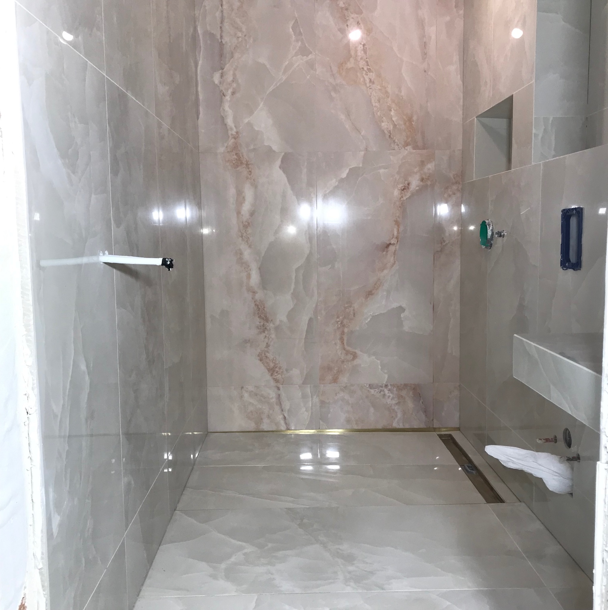 Wykończona łazienka z beżowymi płytkami imitującymi marmur, widoczny odpływ liniowy, półka i wnęka w ścianie. Częściowo zakryta umywalka z baterią.