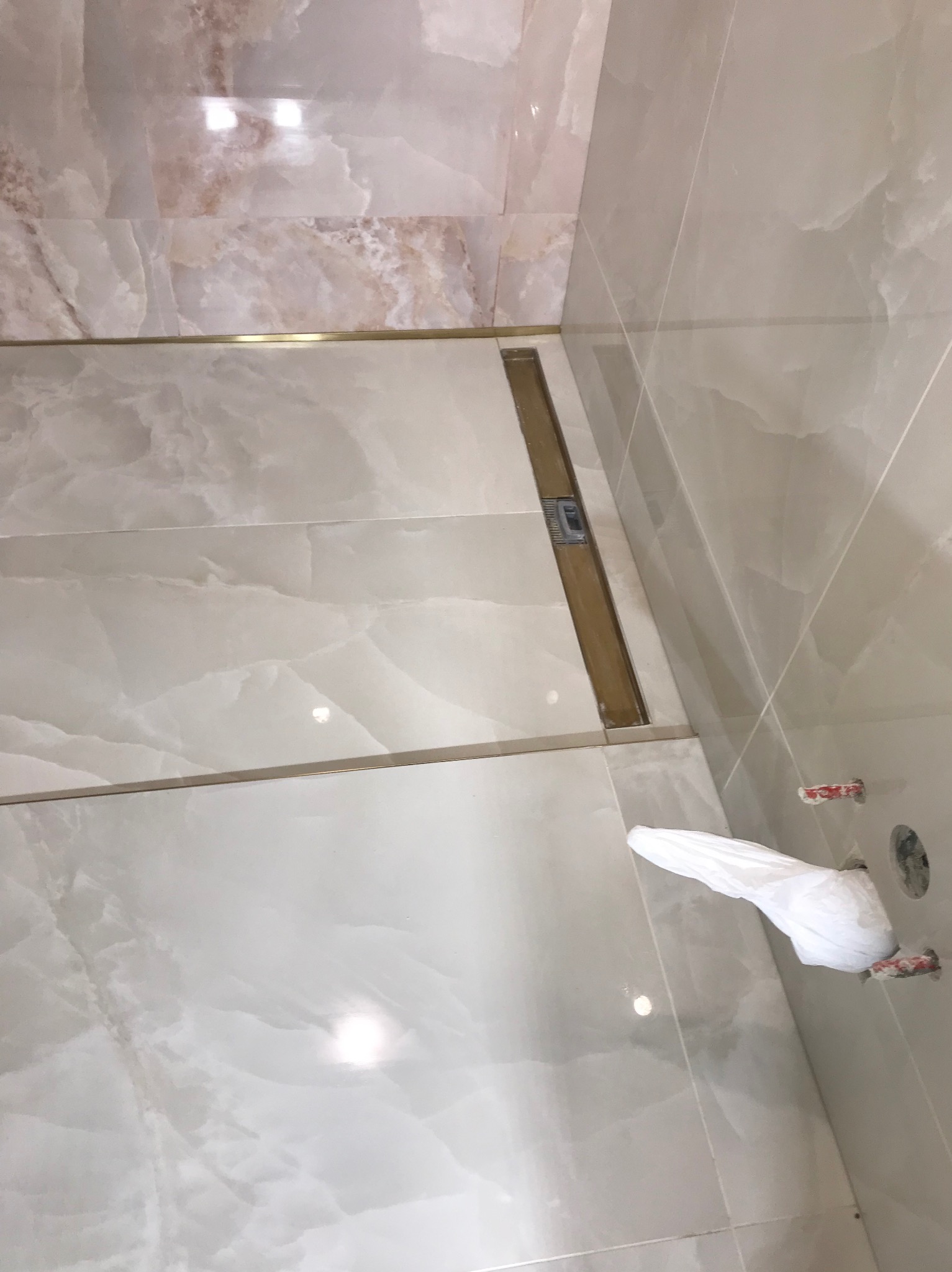 Wykończona łazienka z beżowymi płytkami imitującymi marmur, złotym odpływem liniowym i zabezpieczonymi rurami. Widok z góry, ukośne ujęcie.