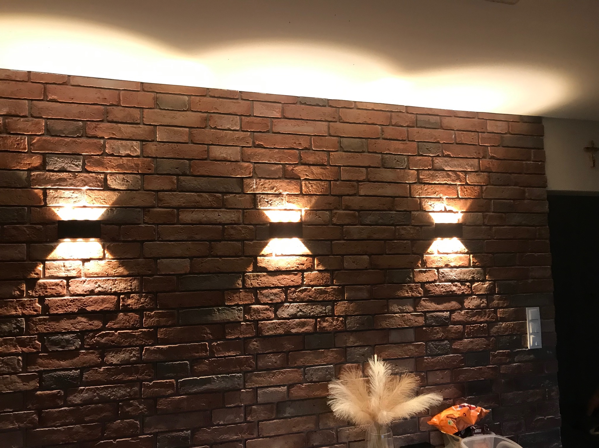 Ściana z cegły klinkierowej oświetlona trzema lampami ściennymi, tworzącymi dekoracyjny efekt światłocienia. W tle widoczny fragment sufitu i dekoracje.