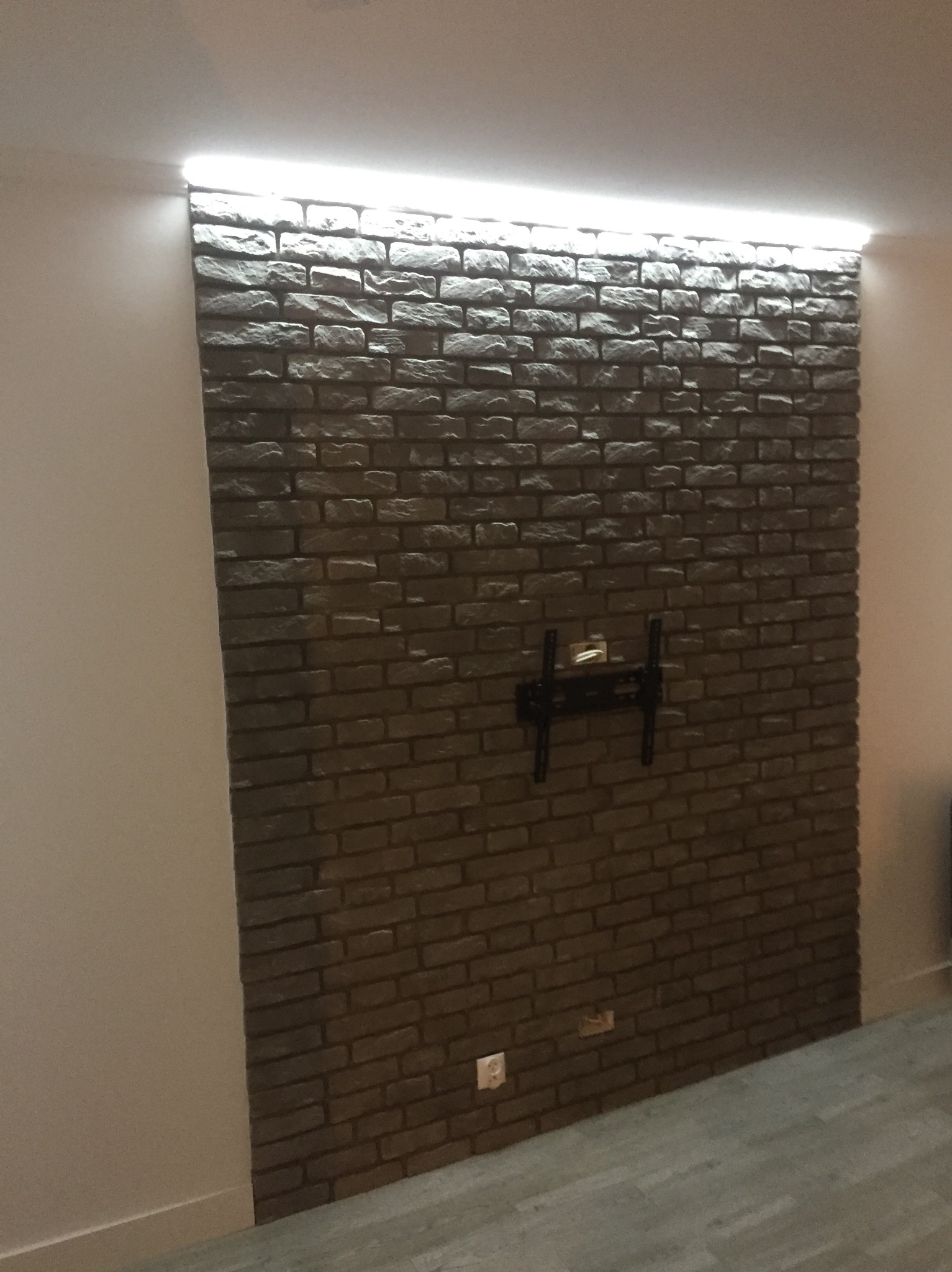 Ściana z cegły dekoracyjnej z podświetleniem LED u góry i uchwytem na telewizor. Widoczny fragment podłogi i ściany bocznej. Gniazdko elektryczne na dole ściany.