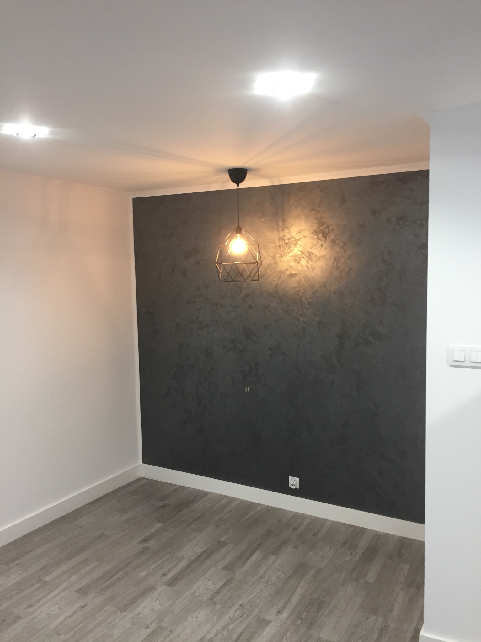 Pokój po remoncie: szara ściana z efektem dekoracyjnym, drewnopodobna podłoga, nowoczesna lampa wisząca. Widoczne gniazdko elektryczne i listwy przypodłogowe.