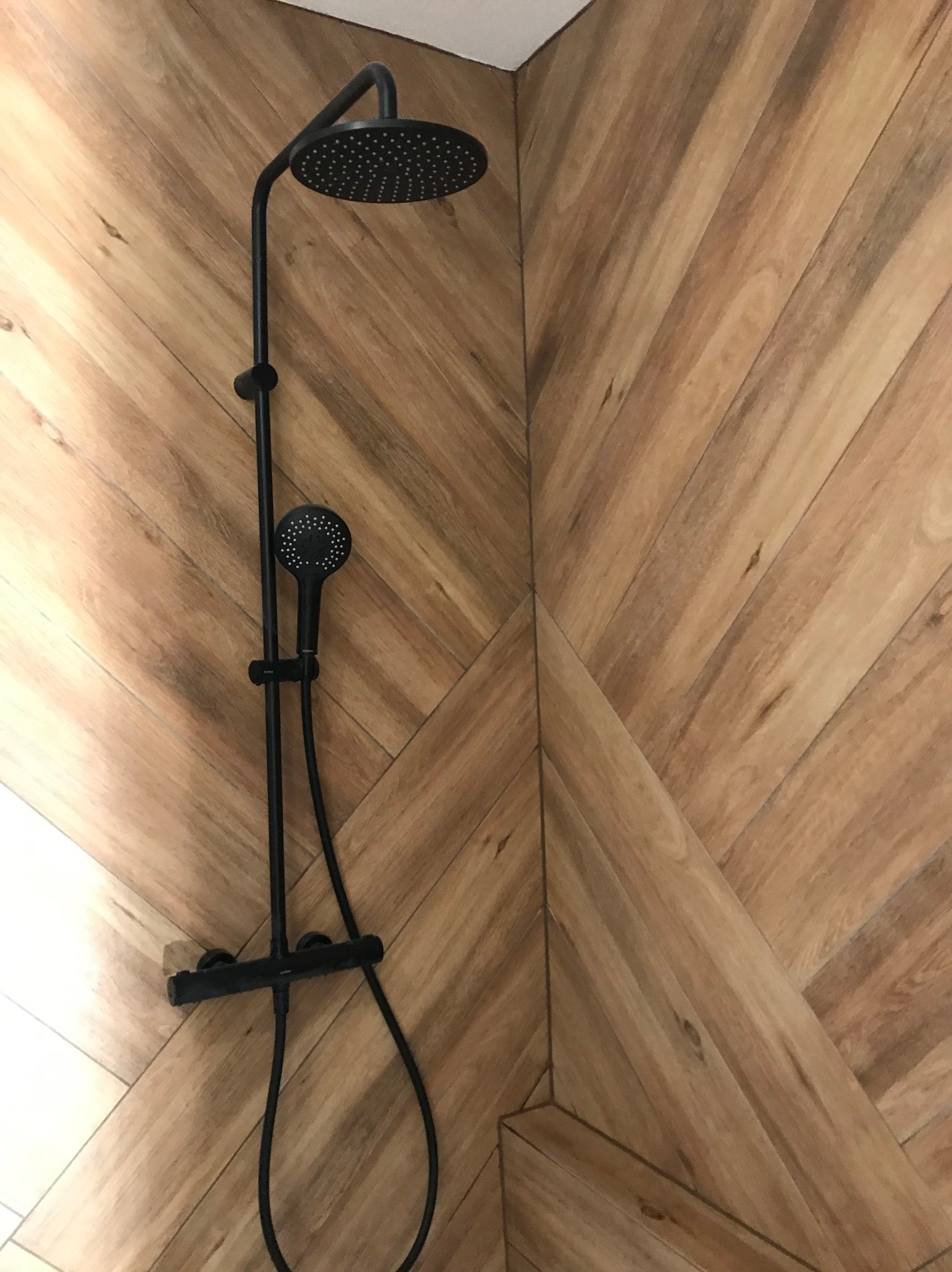 Nowoczesny zestaw prysznicowy w kolorze czarnym, zamontowany na ścianie wyłożonej płytkami imitującymi drewno ułożonymi w jodełkę. Minimalistyczny design.