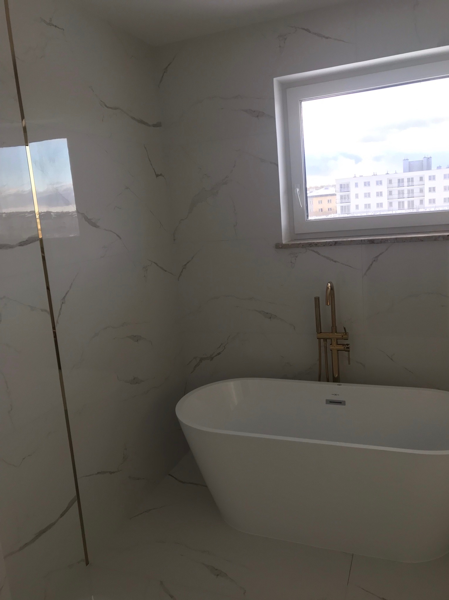 Biała wanna wolnostojąca z złotą armaturą na tle ściany wyłożonej płytkami imitującymi marmur w łazience z oknem. Widok na budynki za oknem.