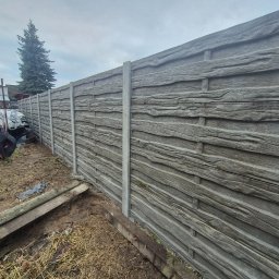 Drew-Pan Ogrodzenia - Długi, szary betonowy płot imitujący drewno, z betonowymi słupkami, oddzielający posesję. W tle budynek i wysokie drzewo iglaste.