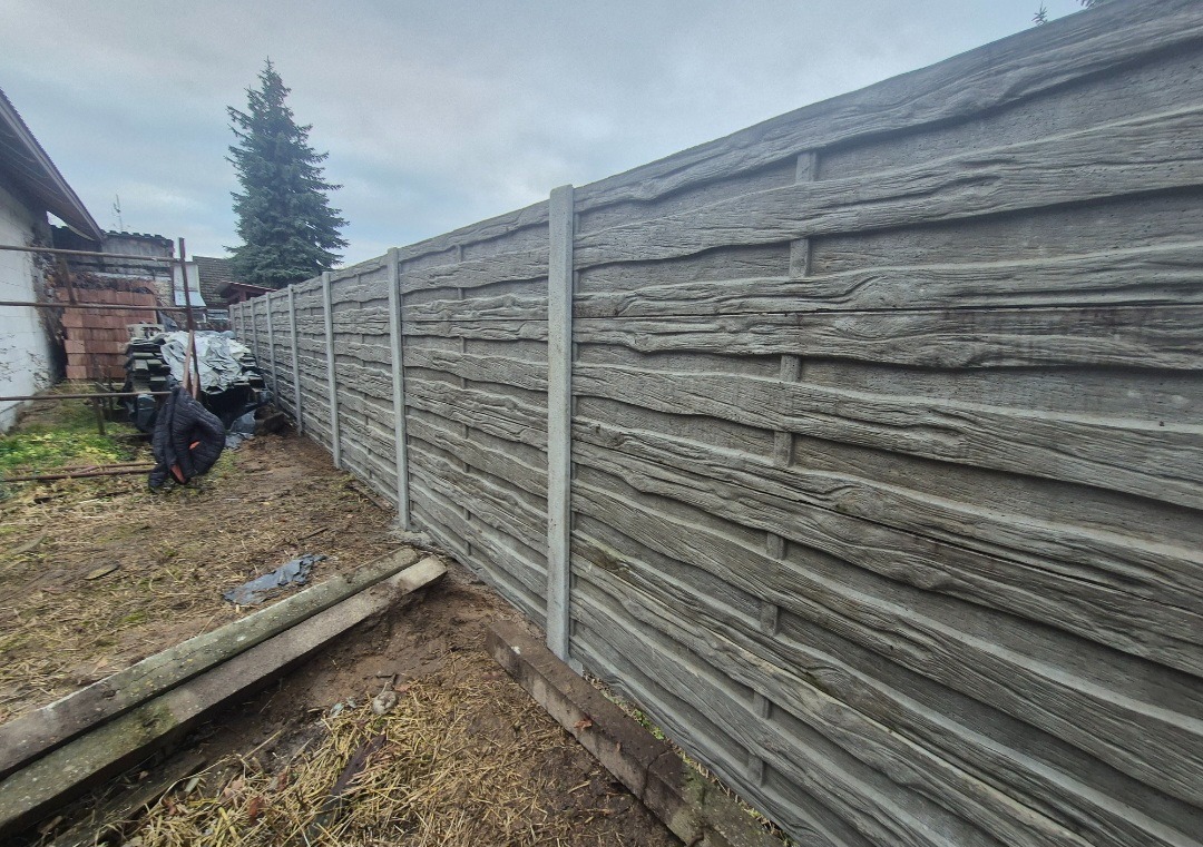 Długi, szary betonowy płot imitujący drewno, z betonowymi słupkami, oddzielający posesję. W tle budynek i wysokie drzewo iglaste.