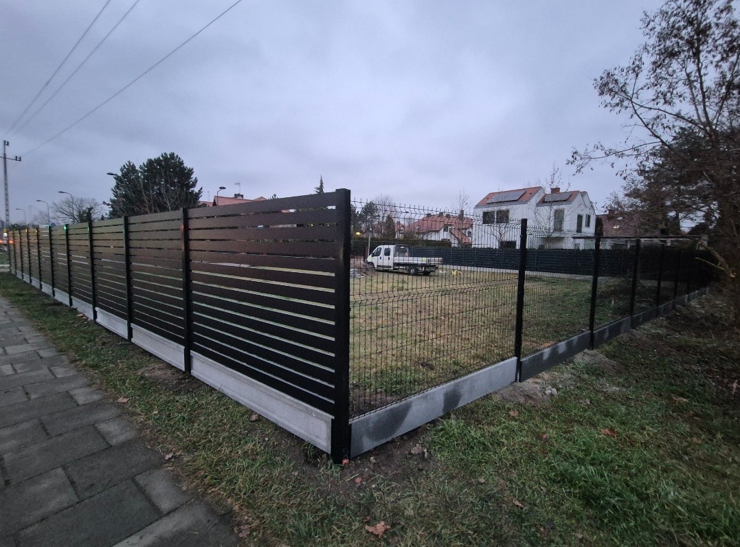 Nowoczesne ogrodzenie: poziome, czarne panele na betonowej podmurówce, siatka z tyłu. Widok z boku, w tle domy i pochmurne niebo.