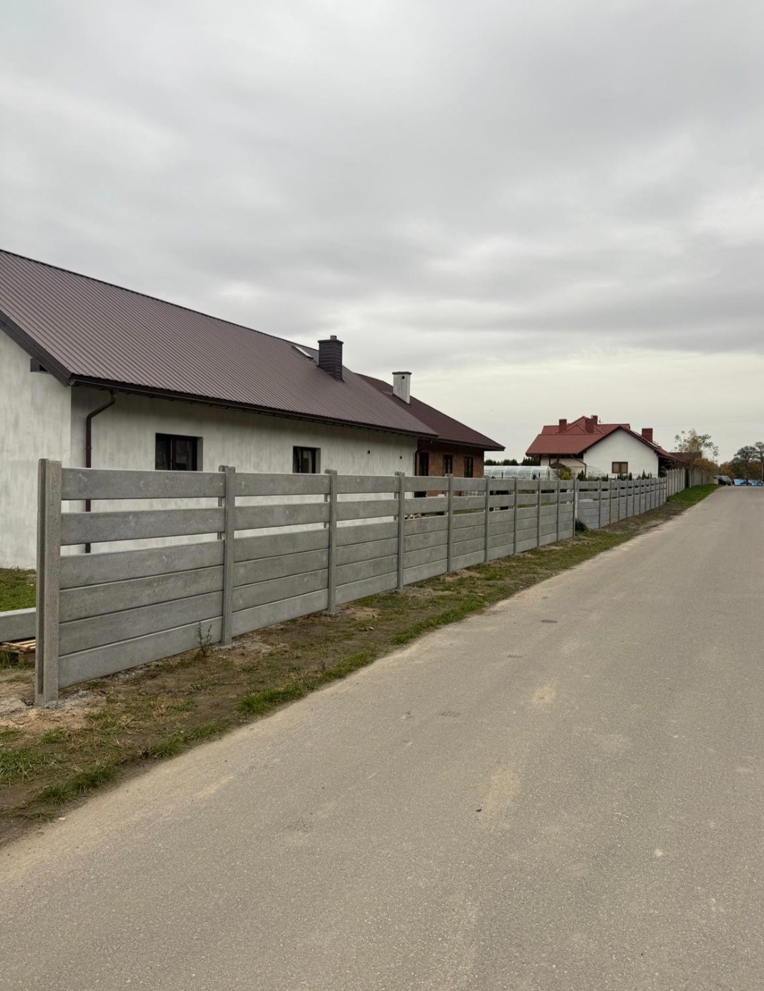 Nowoczesne ogrodzenie betonowe z poziomymi panelami, oddzielające posesję od drogi. Widok z perspektywy ulicy, domy w tle, pochmurne niebo.