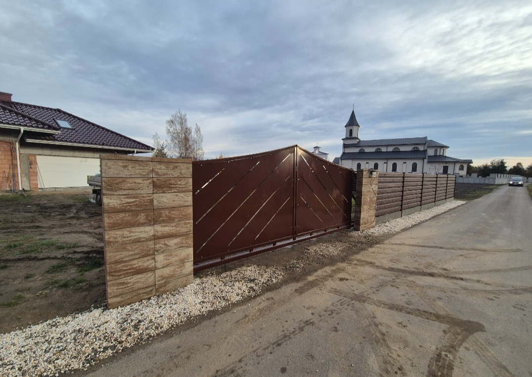 Nowoczesna brama przesuwna z metalu w kolorze brązowym, z ukośnymi wycięciami, osadzona na słupkach z kamienia łupanego. W tle kościół i ogrodzenie panelowe.