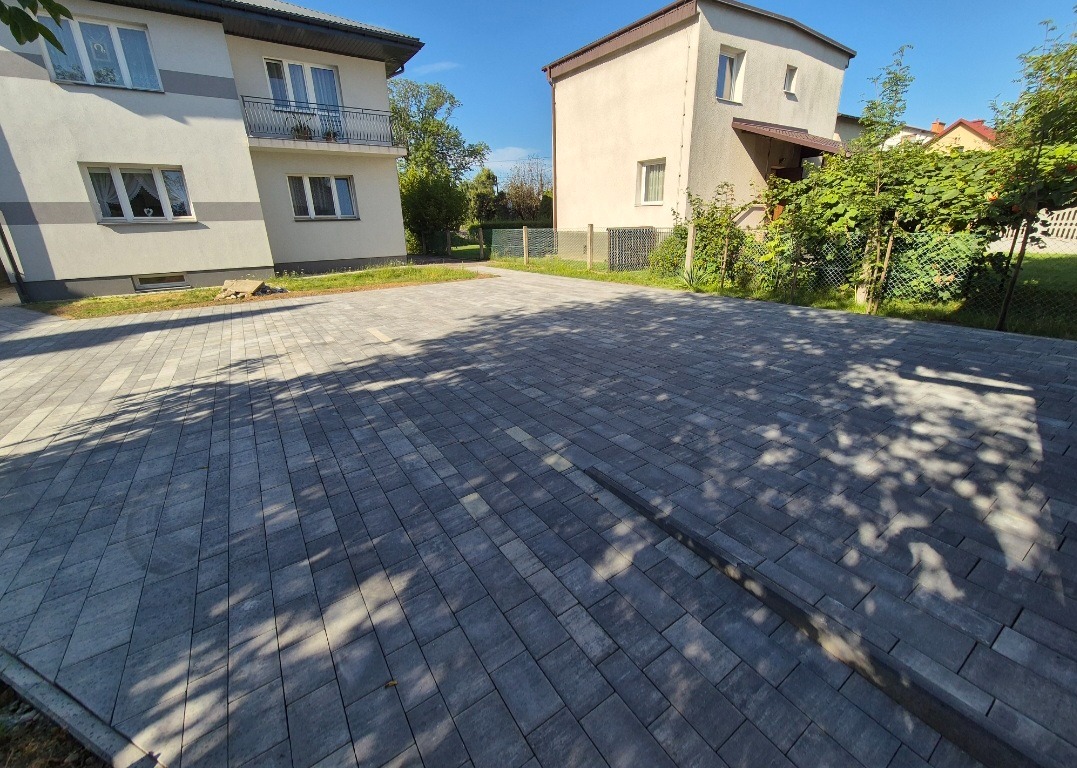 Ułożona szara kostka brukowa na podjeździe między dwoma budynkami, z widocznym fragmentem ogrodzenia w tle. Cień drzew na powierzchni kostki.