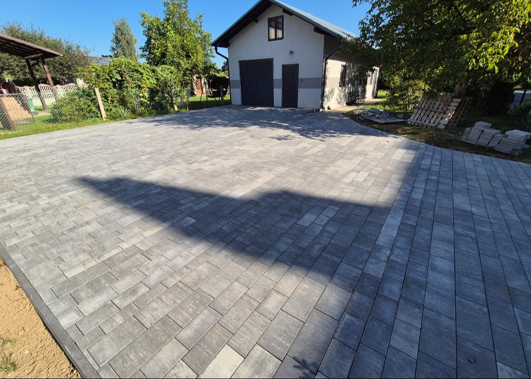 Ułożona kostka brukowa w odcieniach szarości tworzy duży podjazd przed budynkiem z garażem. Widoczne palety z kostką i zieleń w tle.
