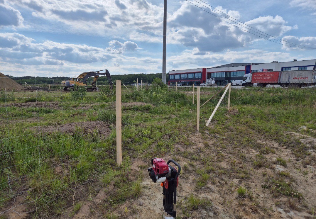 Wbijanie słupków ogrodzeniowych na nierównym terenie z użyciem palownicy spalinowej. W tle widoczny budynek przemysłowy i koparka.