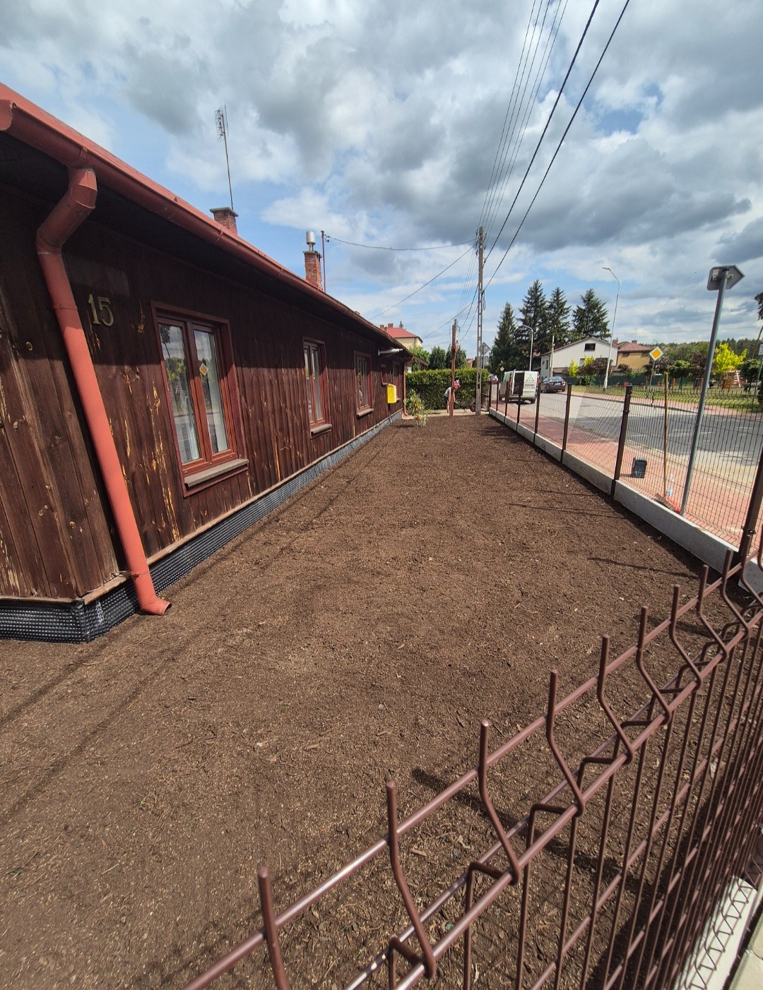 Przygotowany teren pod trawnik, widok zza brązowego ogrodzenia panelowego. Dom z drewnianą elewacją w tle, numer 15 na ścianie. Słoneczny dzień.