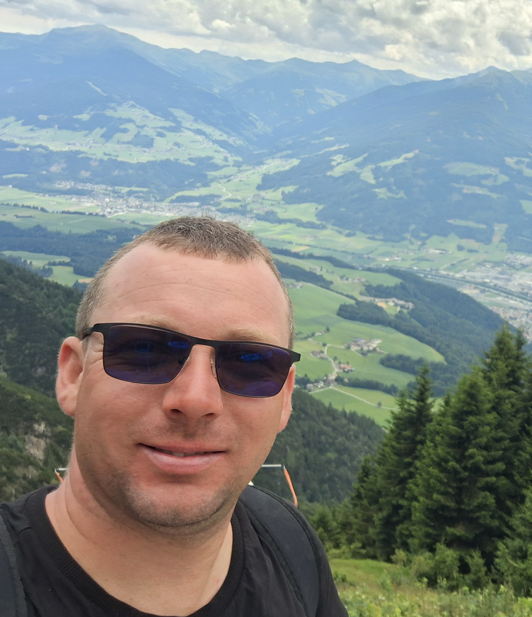 Uśmiechnięty mężczyzna w okularach na tle górskiego krajobrazu z widocznymi dolinami i zabudowaniami. W tle pochmurne niebo.