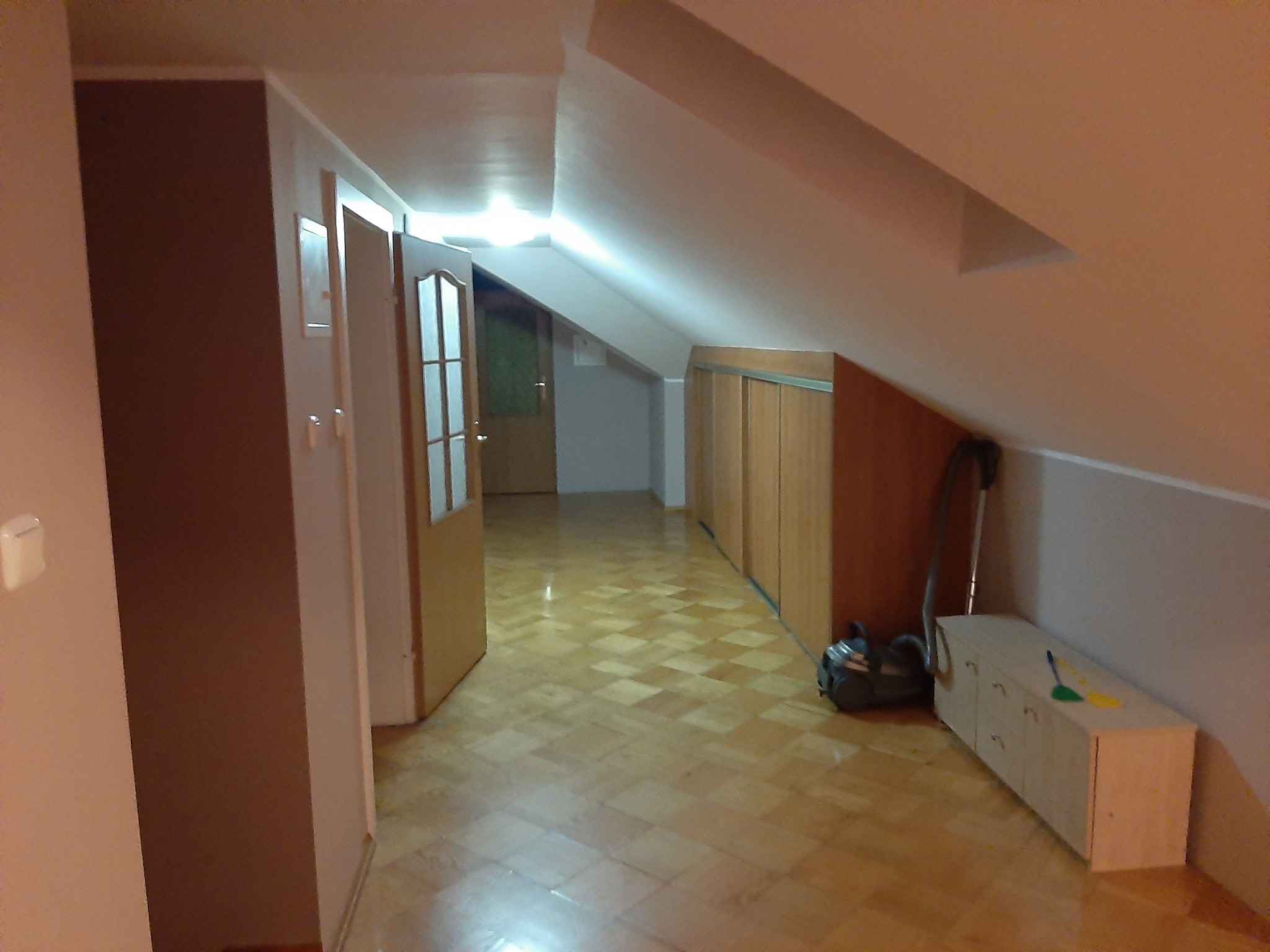 Poddasze po malowaniu: świeżo pomalowane ściany w odcieniach brązu i beżu, parkiet w jodełkę, widoczny odkurzacz i szafa wnękowa. Czyste, jasne wnętrze.