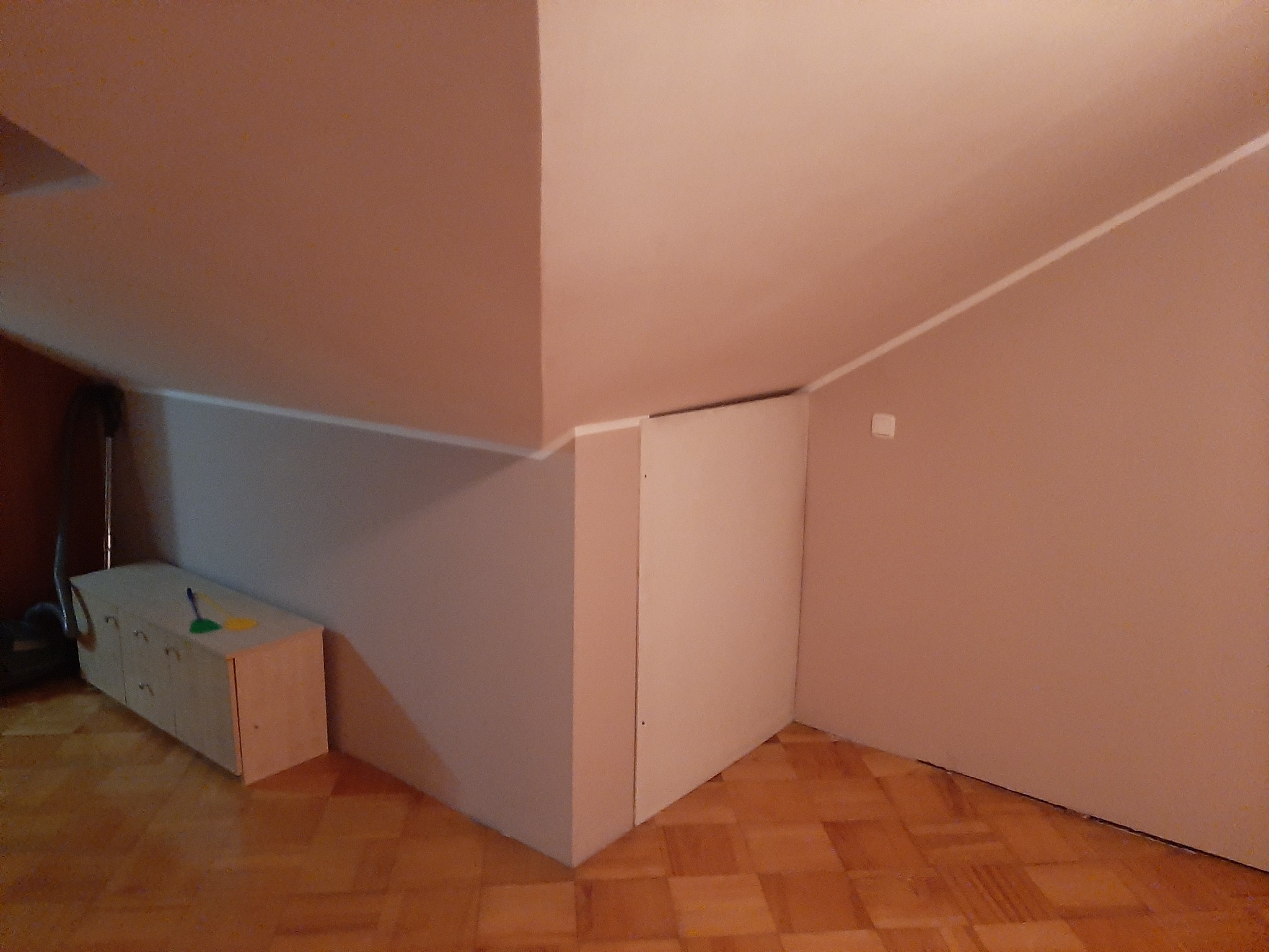 Pomieszczenie na poddaszu po malowaniu: jasne ściany, drewniana podłoga, zabudowa w rogu. Widoczny fragment odkurzacza i szafka z akcesoriami do sprzątania.