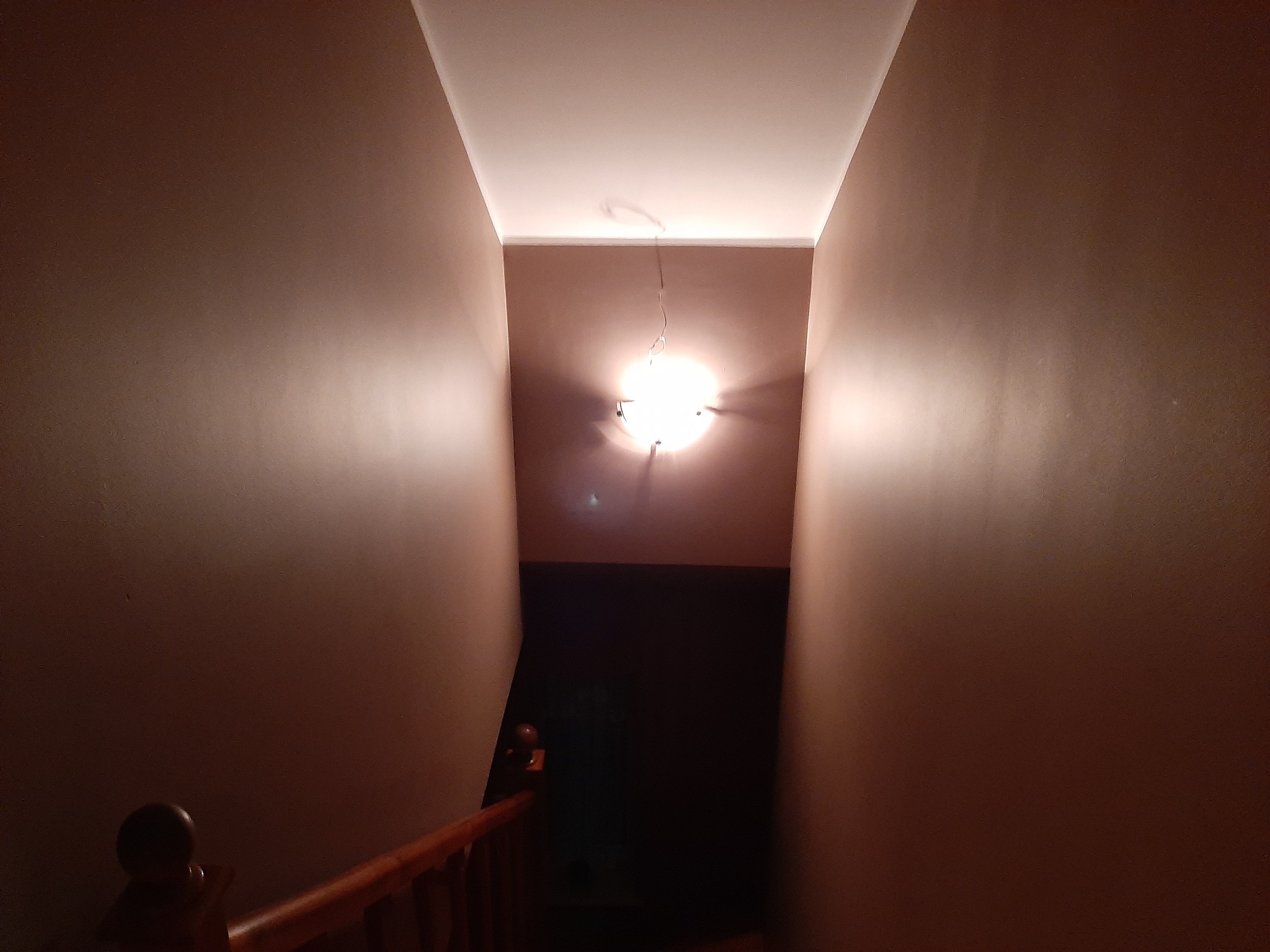 Klatka schodowa z brązowymi ścianami oświetlona przez lampę sufitową. Widok z góry na dół schodów. Drewniana balustrada po lewej stronie.