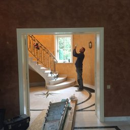 Grzegorz Sasinowski - Wnętrze z marmurową podłogą i schodami, widoczne przez drzwi. Mężczyzna robi zdjęcie smartfonem. Ściany w odcieniach brązu i pomarańczy, dekoracyjna sztukateria.