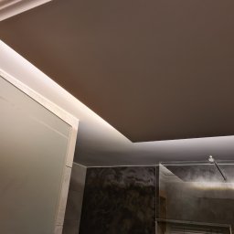 Grzegorz Sasinowski - Nowoczesna łazienka z podwieszanym sufitem i oświetleniem LED. Widoczne szklane elementy kabiny prysznicowej i ciemne płytki imitujące marmur.