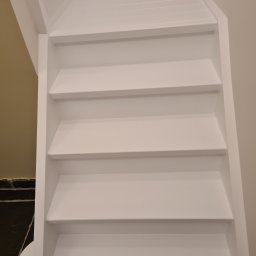 Grzegorz Sasinowski - Białe, odnowione schody drewniane w domu, widok z dołu. Świeżo pomalowane stopnie i boki schodów, kontrastujące z ciemną podłogą i jasnymi ścianami.