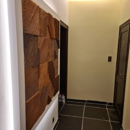 Grzegorz Sasinowski - Korytarz z dekoracyjną, drewnianą płaskorzeźbą na ścianie, podświetloną listwą LED. Ciemna podłoga z płytek, drzwi po prawej stronie. Nowoczesne wykończenie.