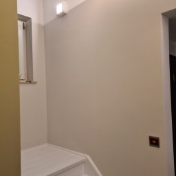 Grzegorz Sasinowski - Jasne wnętrze po odnowieniu: białe schody, kremowe ściany, nowoczesne oświetlenie i minimalistyczny włącznik. Widok z dołu, podkreślający świeżość i prostotę.