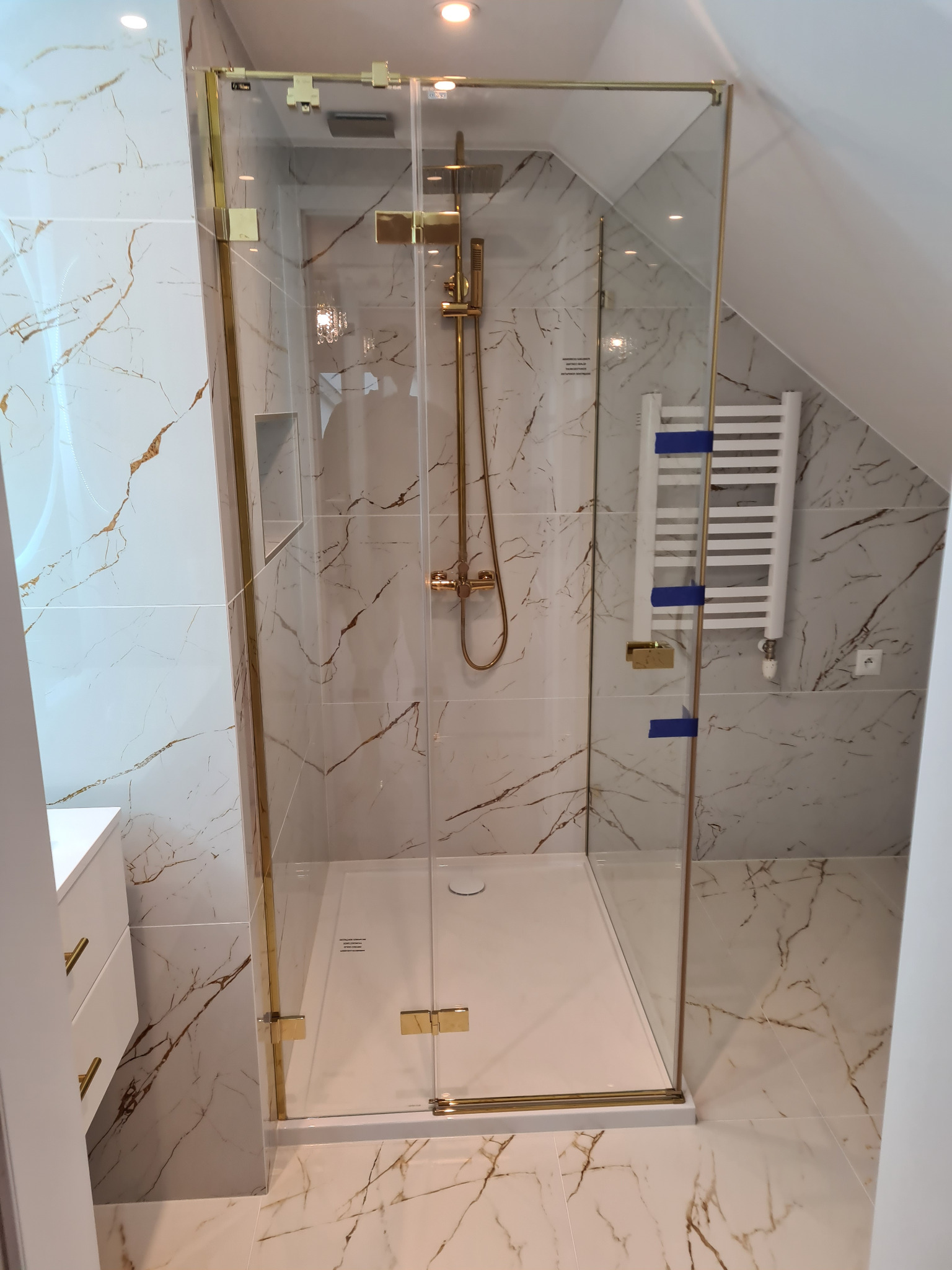 Nowoczesna kabina prysznicowa z złotymi detalami w łazience na poddaszu, wykończonej marmurowymi płytkami z brązowymi żyłami i białym grzejnikiem drabinkowym.