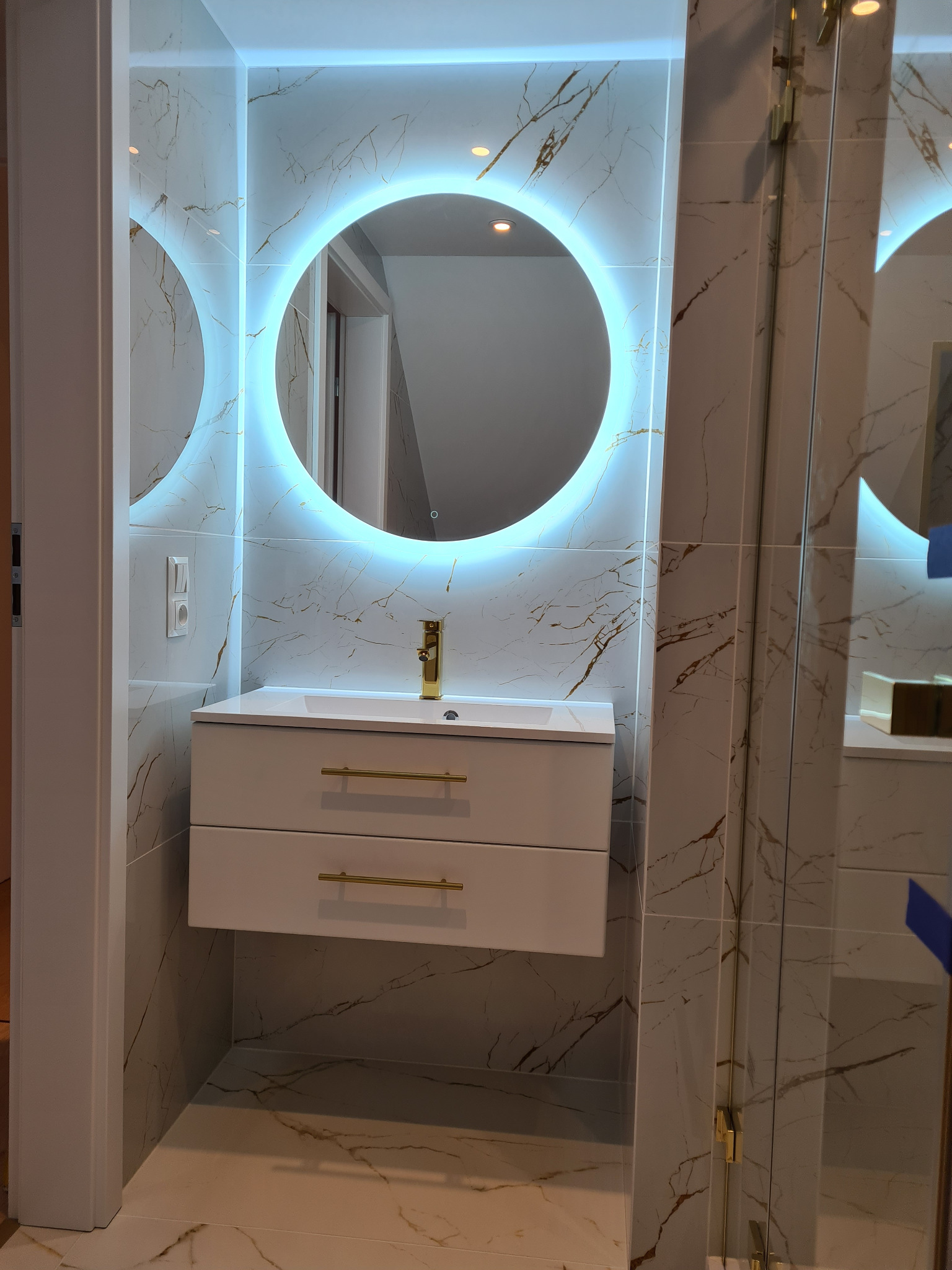 Nowoczesna łazienka z marmurowymi płytkami, okrągłym lustrem z podświetleniem LED, białą szafką pod umywalką z złotymi uchwytami i złotą armaturą.