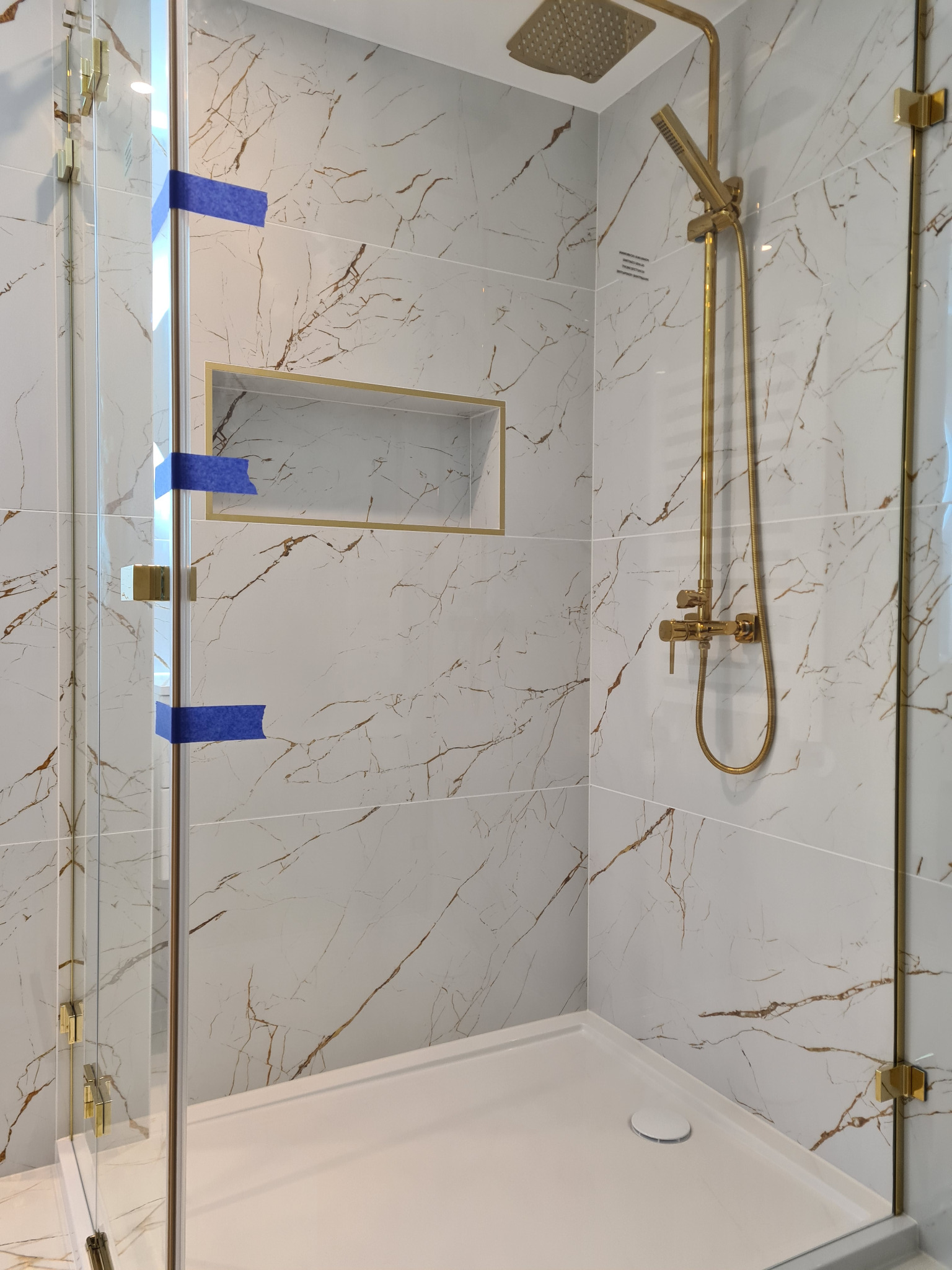 Wykończona kabina prysznicowa z białymi płytkami imitującymi marmur z złotymi akcentami, złotą armaturą i wnęką. Na szkle niebieska taśma zabezpieczająca.