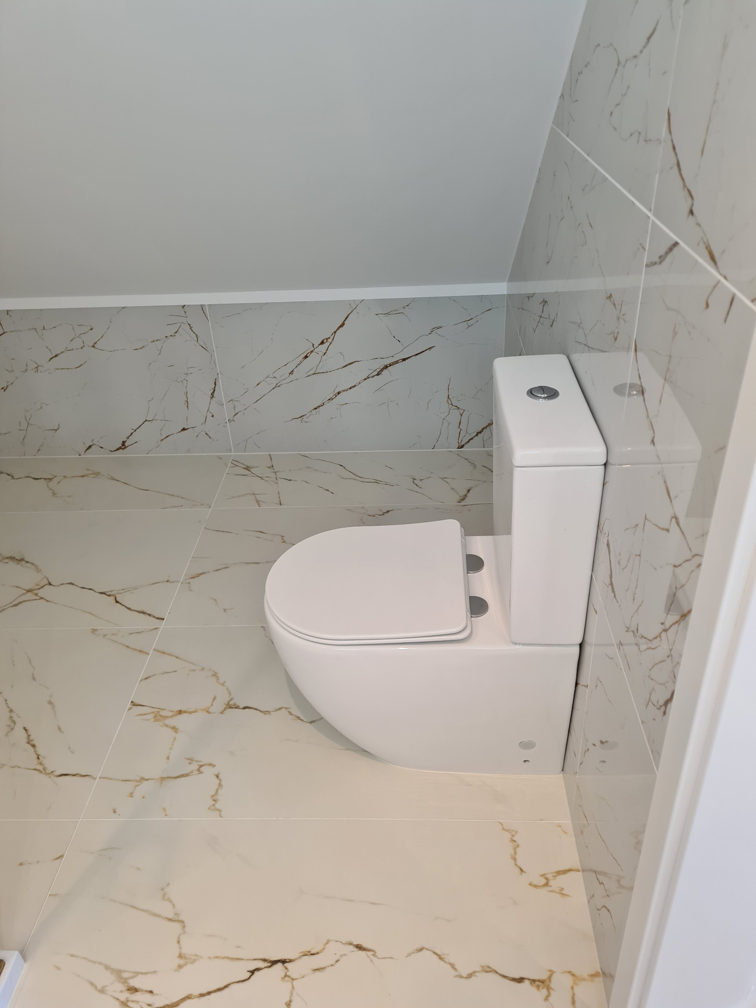 Biała toaleta w wykończonej łazience na poddaszu, z płytkami imitującymi marmur z brązowymi żyłami na ścianach i podłodze. Minimalistyczny styl.