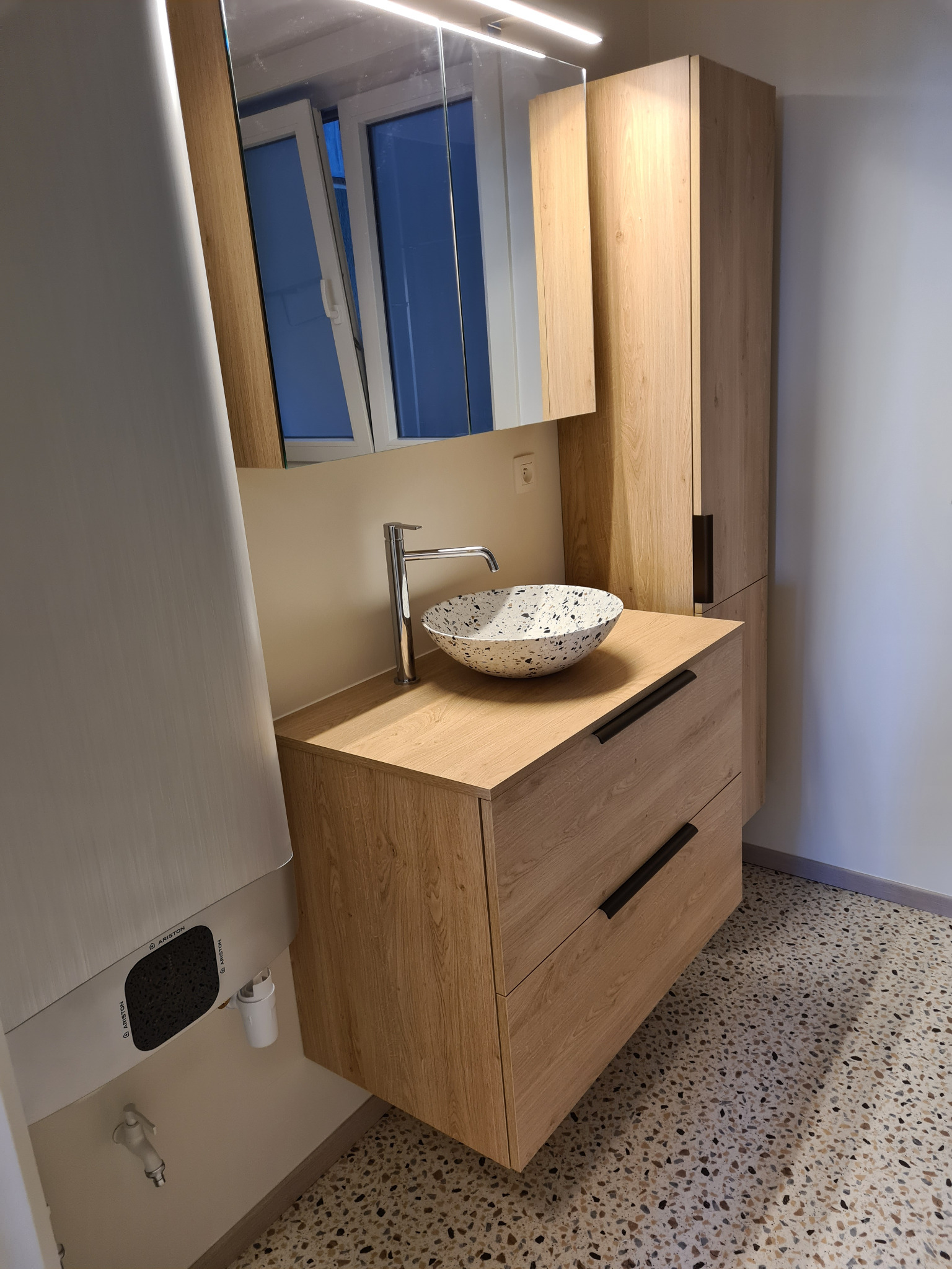 Nowoczesna łazienka z drewnianą szafką pod umywalką w terrazzo, lustrem z oknem w tle i wysoką szafką. Widoczny bojler i podłoga w stylu lastryko.