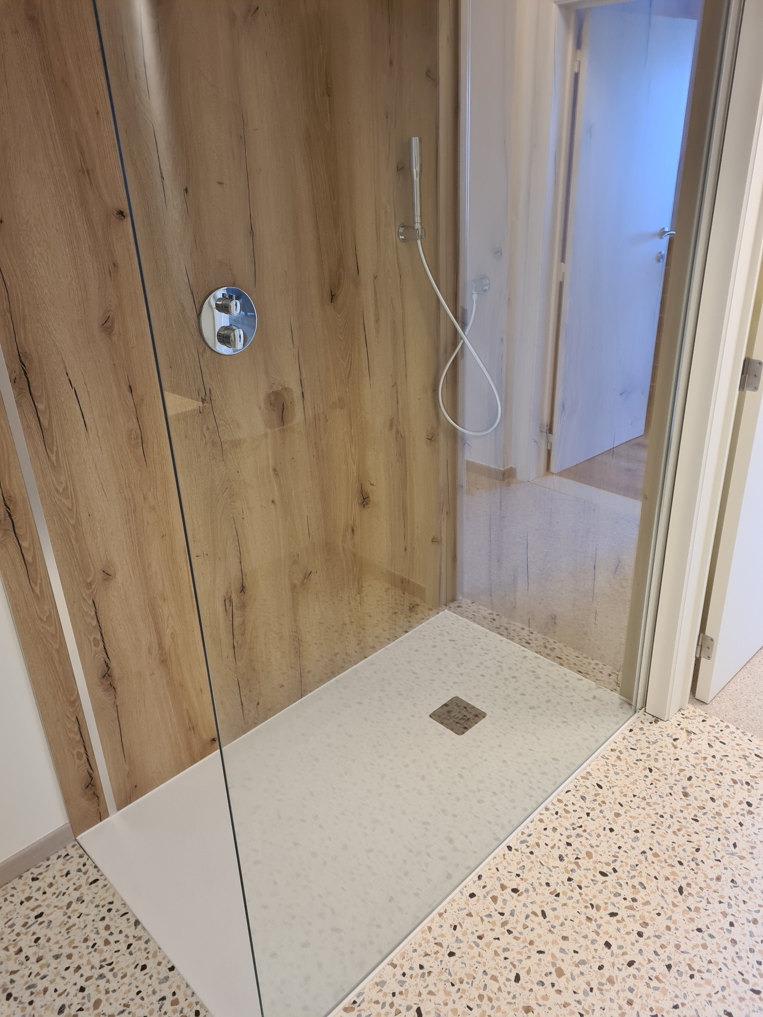 Nowoczesna kabina prysznicowa z drewnopodobną ścianą, szklaną przegrodą i brodzikiem z mozaiką. Widoczny fragment łazienki z terazzo. Minimalistyczny design.