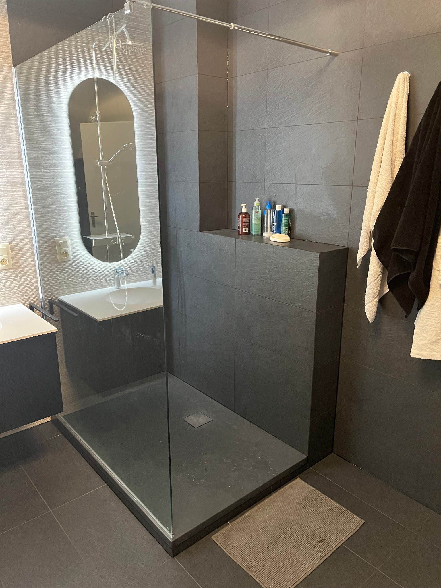 Nowoczesna łazienka z kabiną typu walk-in, wykończona ciemnymi płytkami. Uwagę zwraca podświetlane lustro i minimalistyczny design. Widoczne kosmetyki i ręczniki.