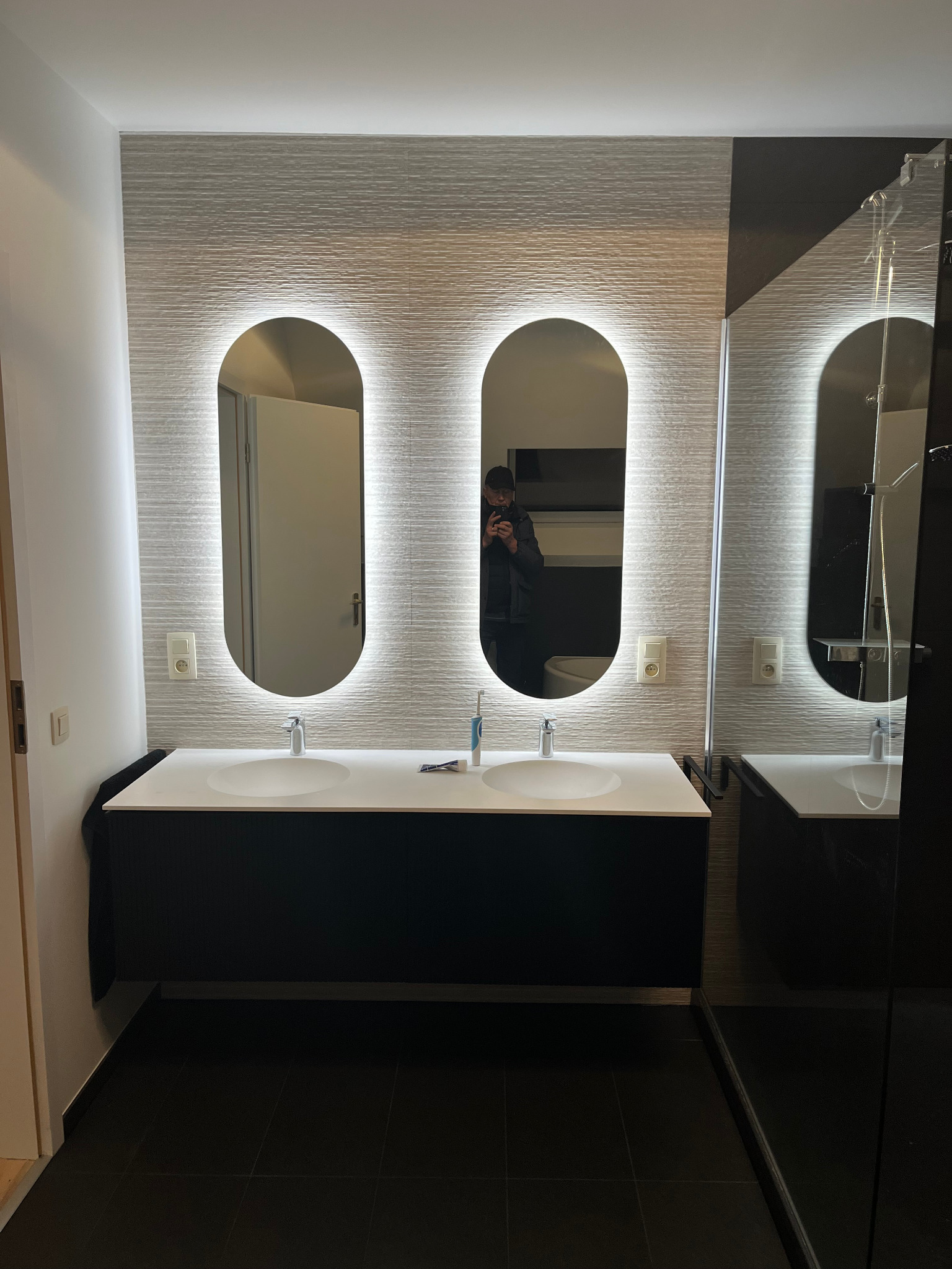 Nowoczesna łazienka z podwójną umywalką, podświetlanymi lustrami i teksturowanymi płytkami ściennymi. Minimalistyczny design z czarnymi akcentami i ciemną podłogą.